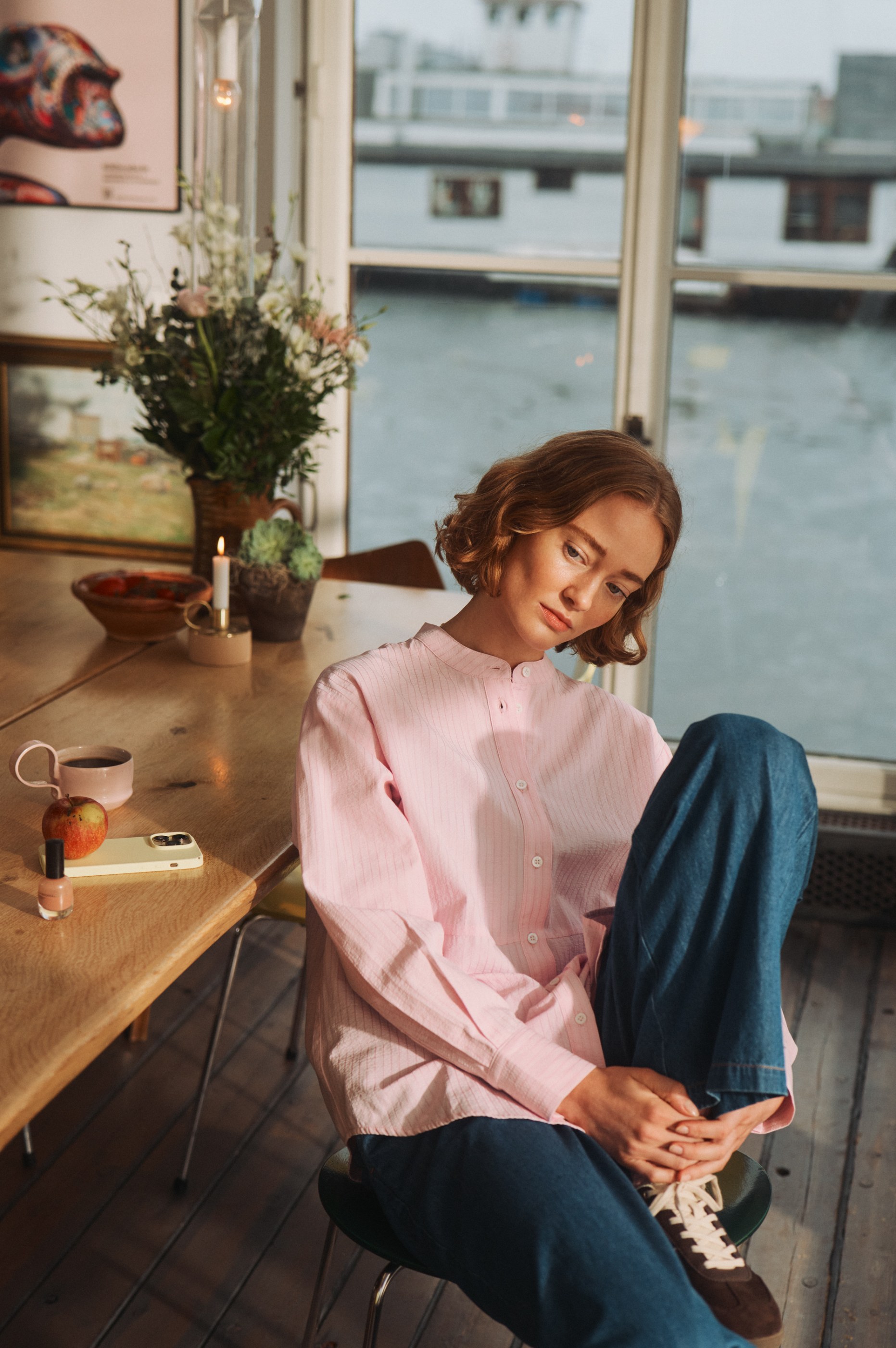 Pensive young woman in a pink striped shirt sits by a window overlooking water.