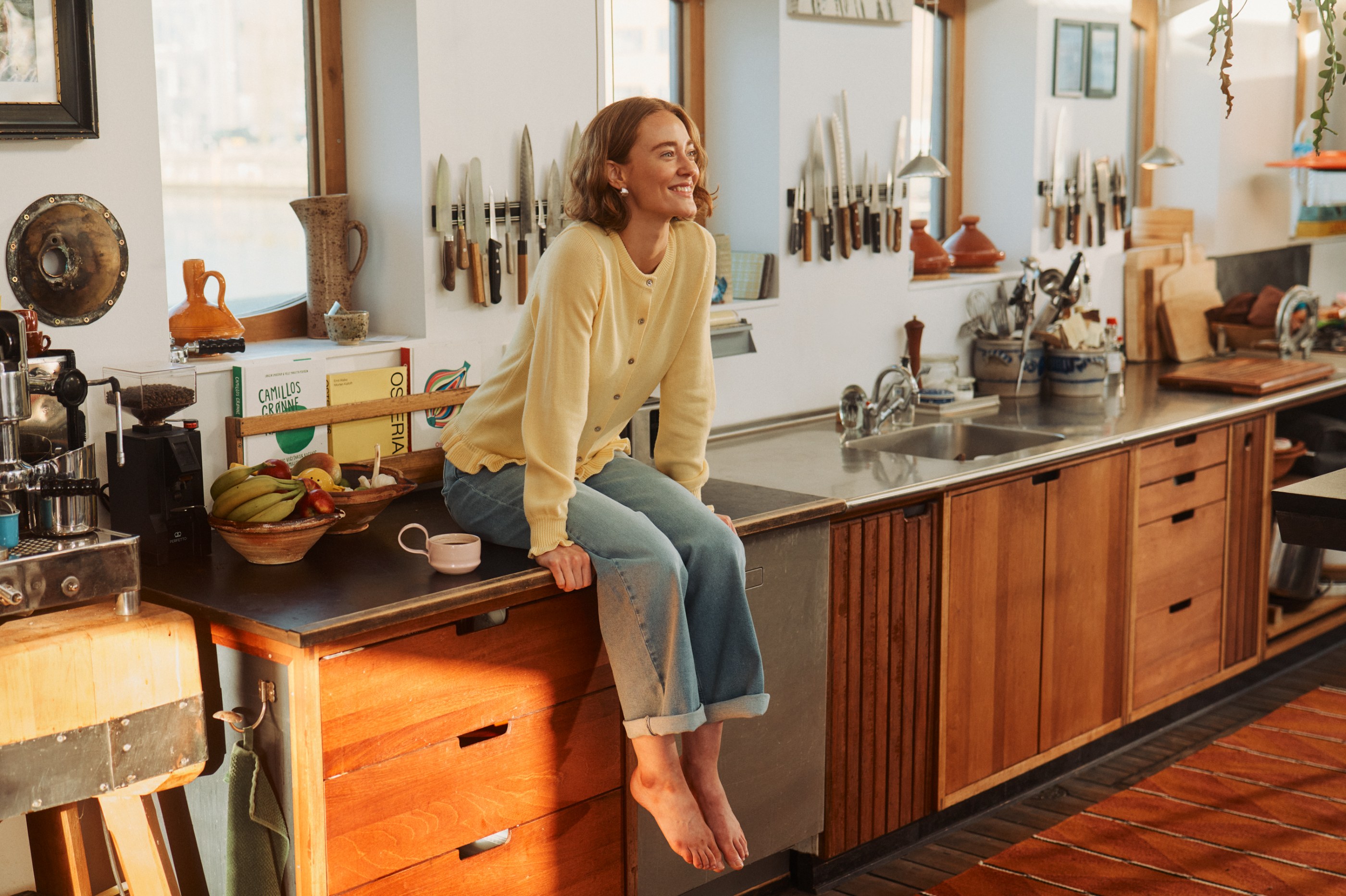 Smiling woman in yellow cardigan and jeans sitting on a rustic kitchen counter.