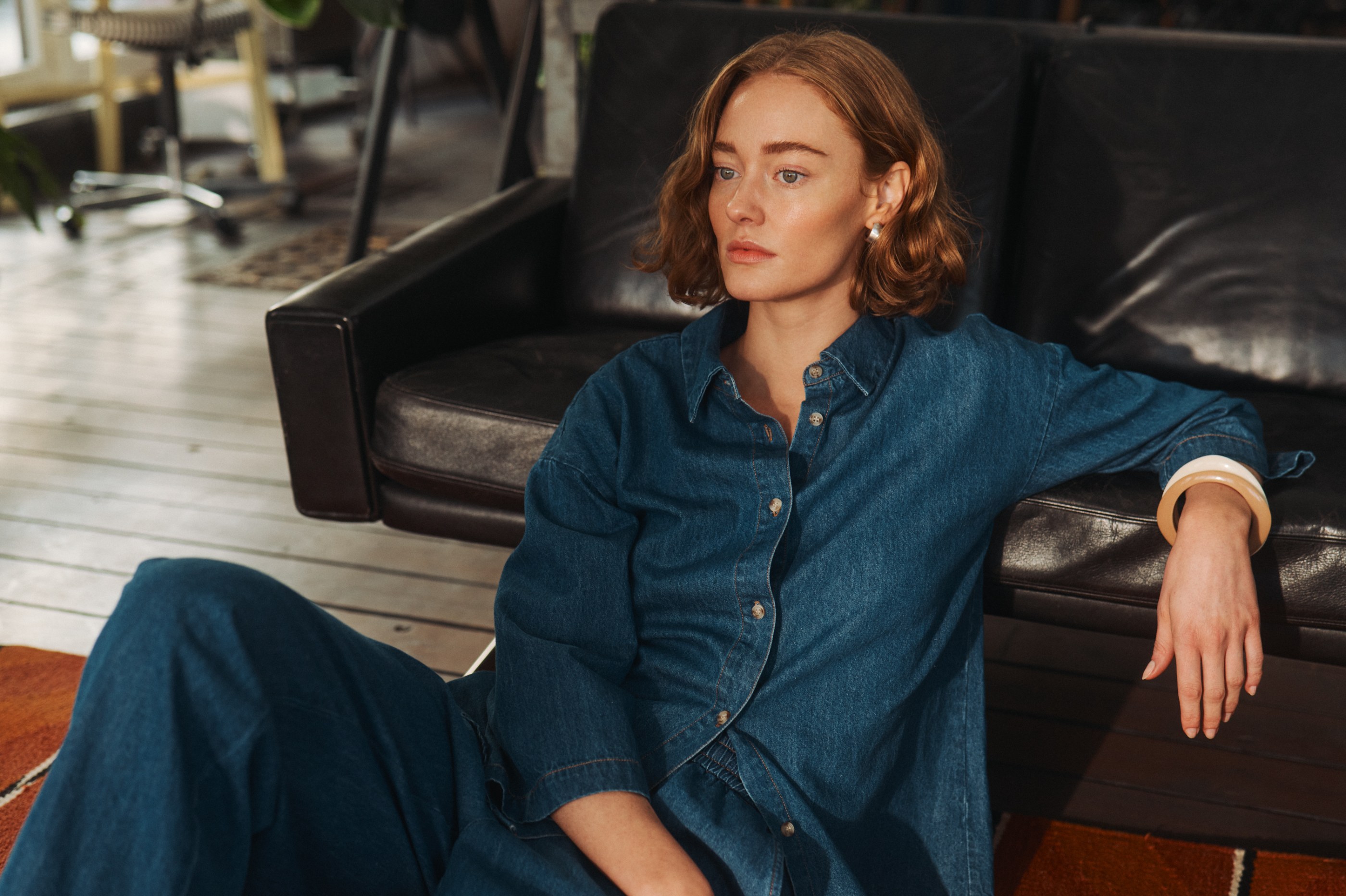 Ginger-haired woman in a denim outfit sitting on the floor against a dark leather couch.
