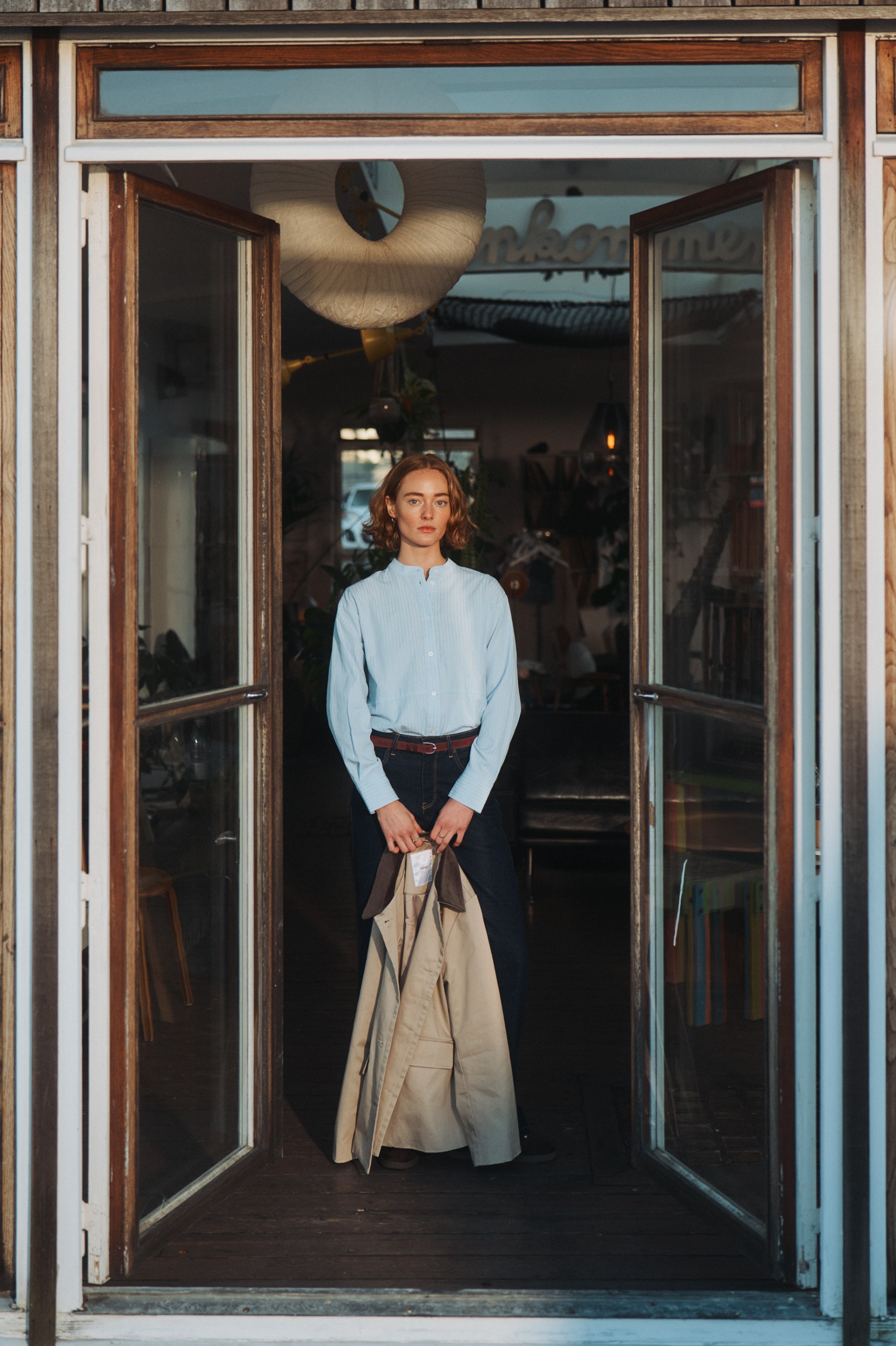Woman in blue shirt and jeans holds a beige trench coat, standing in a rustic doorway.