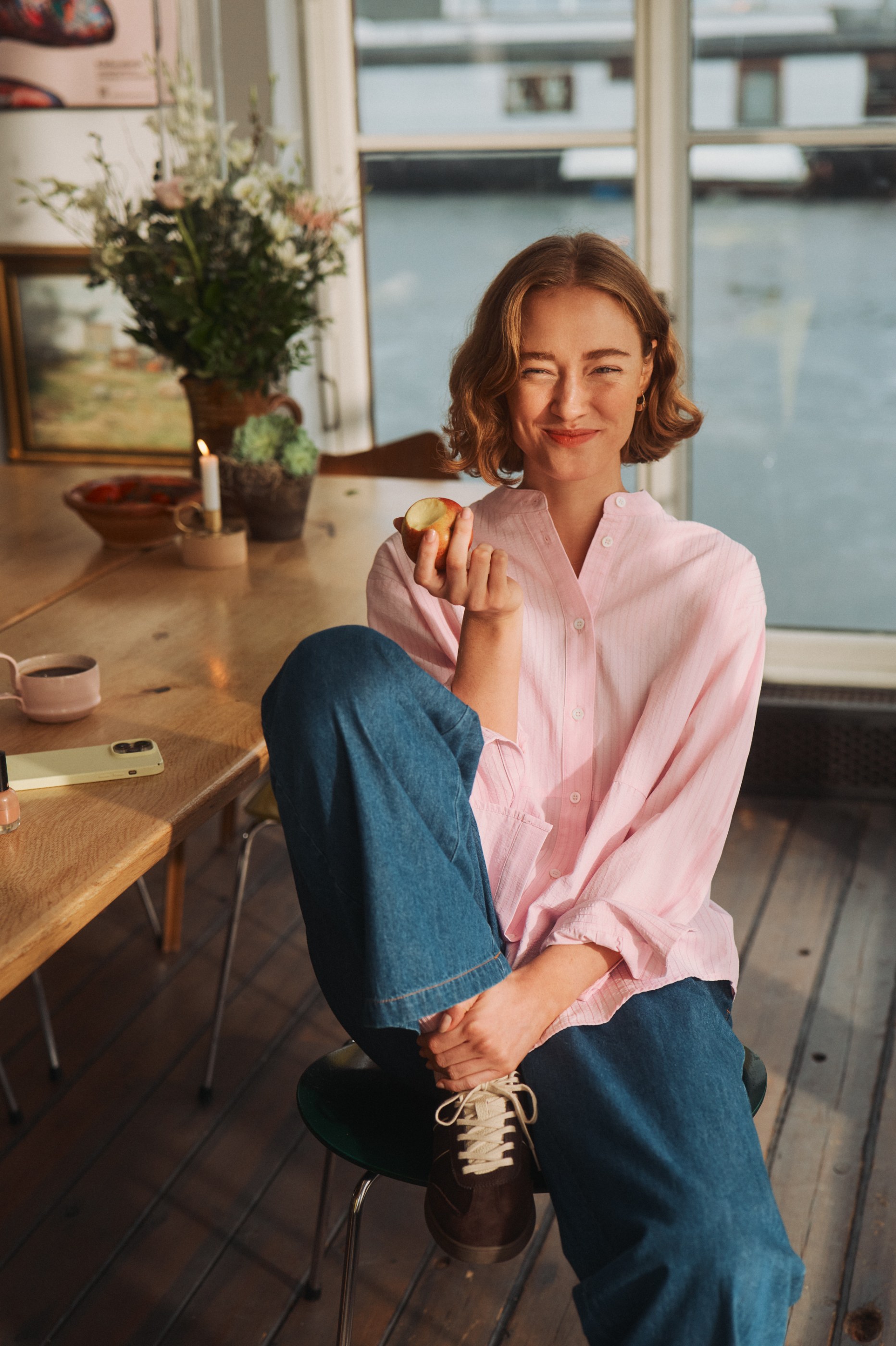 Winking woman in pink shirt and blue jeans holds a half-eaten apple by a window.