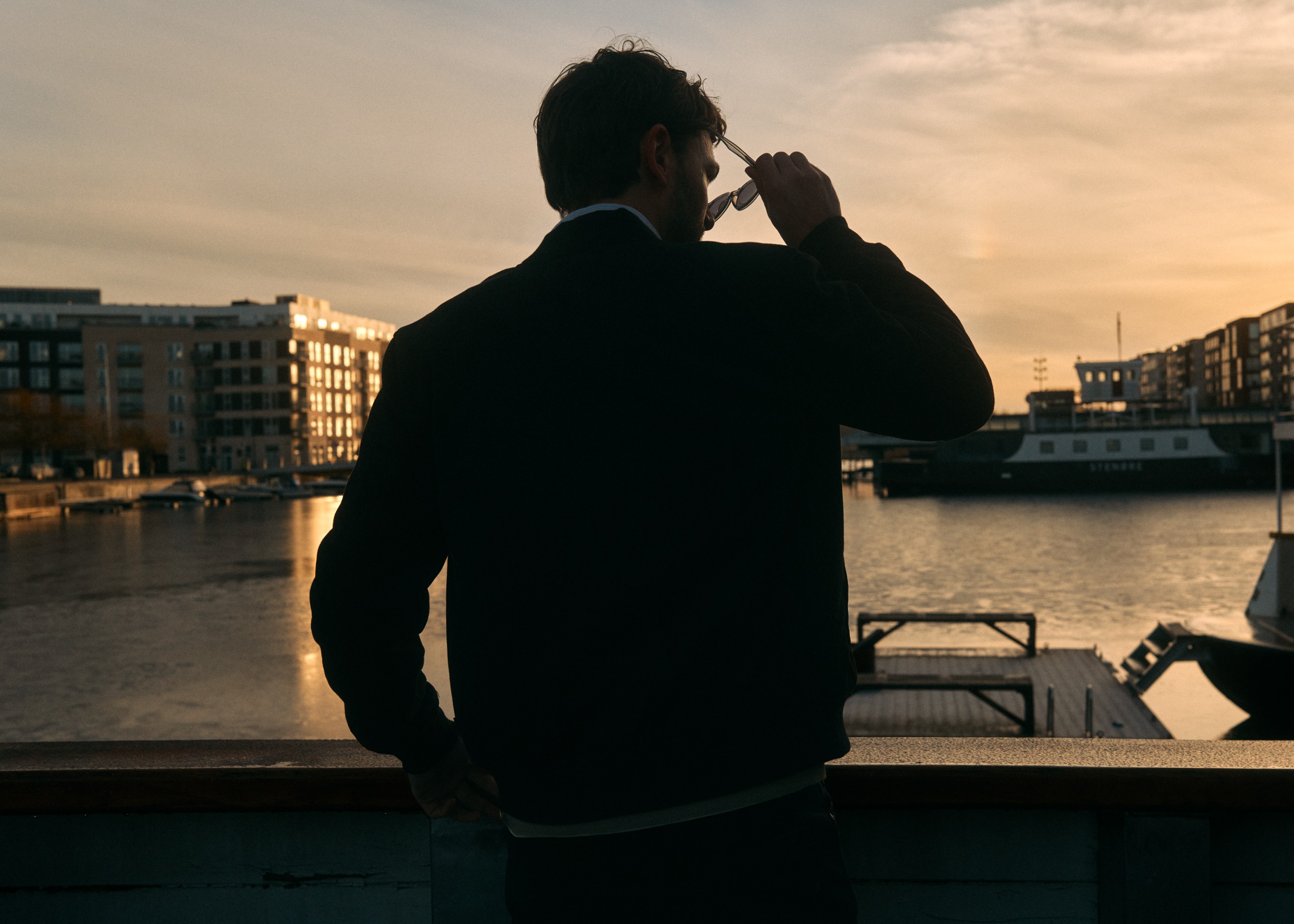 Man in silhouette holds sunglasses, gazing at a golden sunset over a harbor.