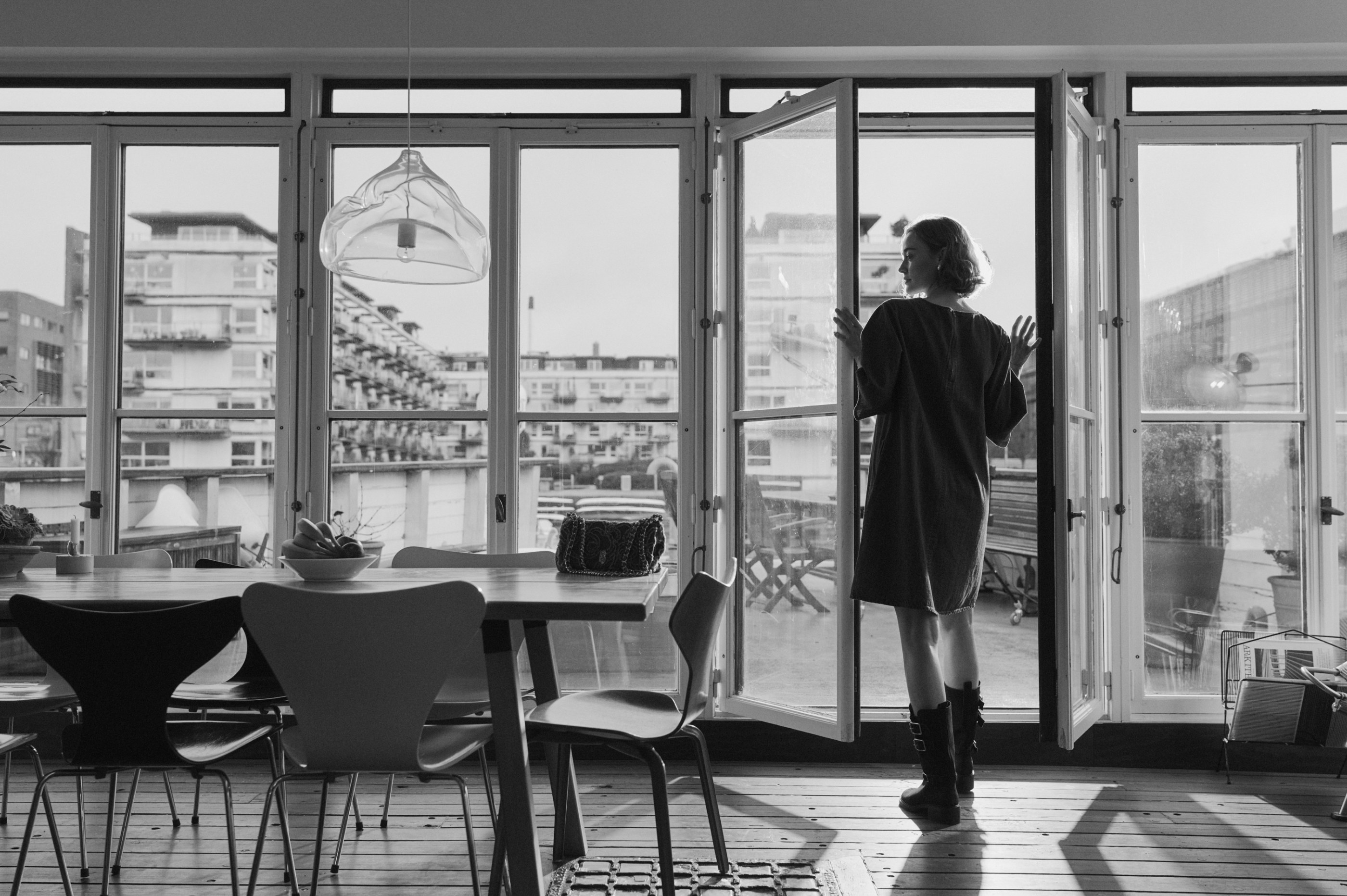 Woman in dress and boots by open balcony door, looking out at city view.