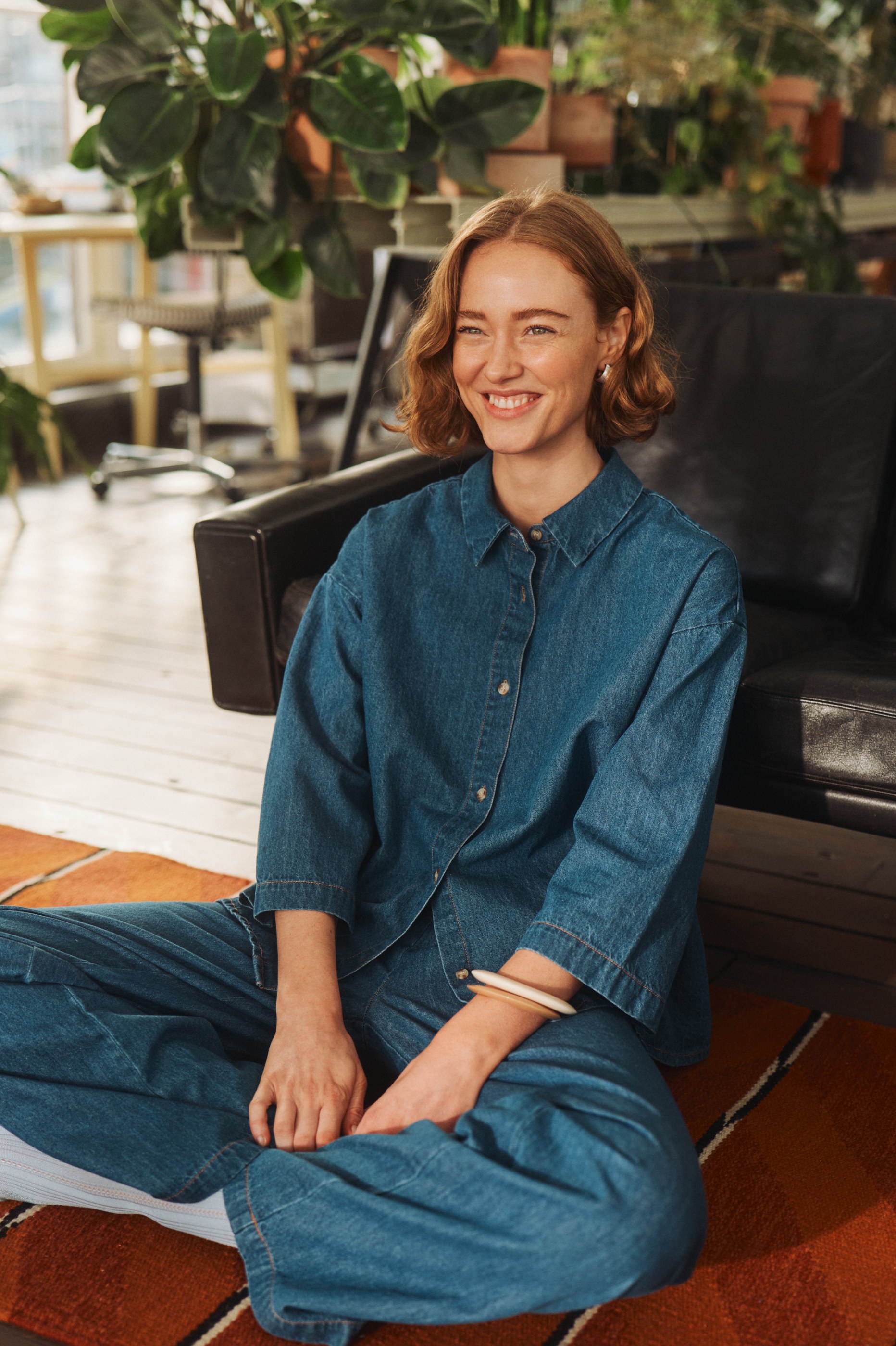 Smiling woman with reddish hair in a denim outfit sitting on a rug surrounded by plants.