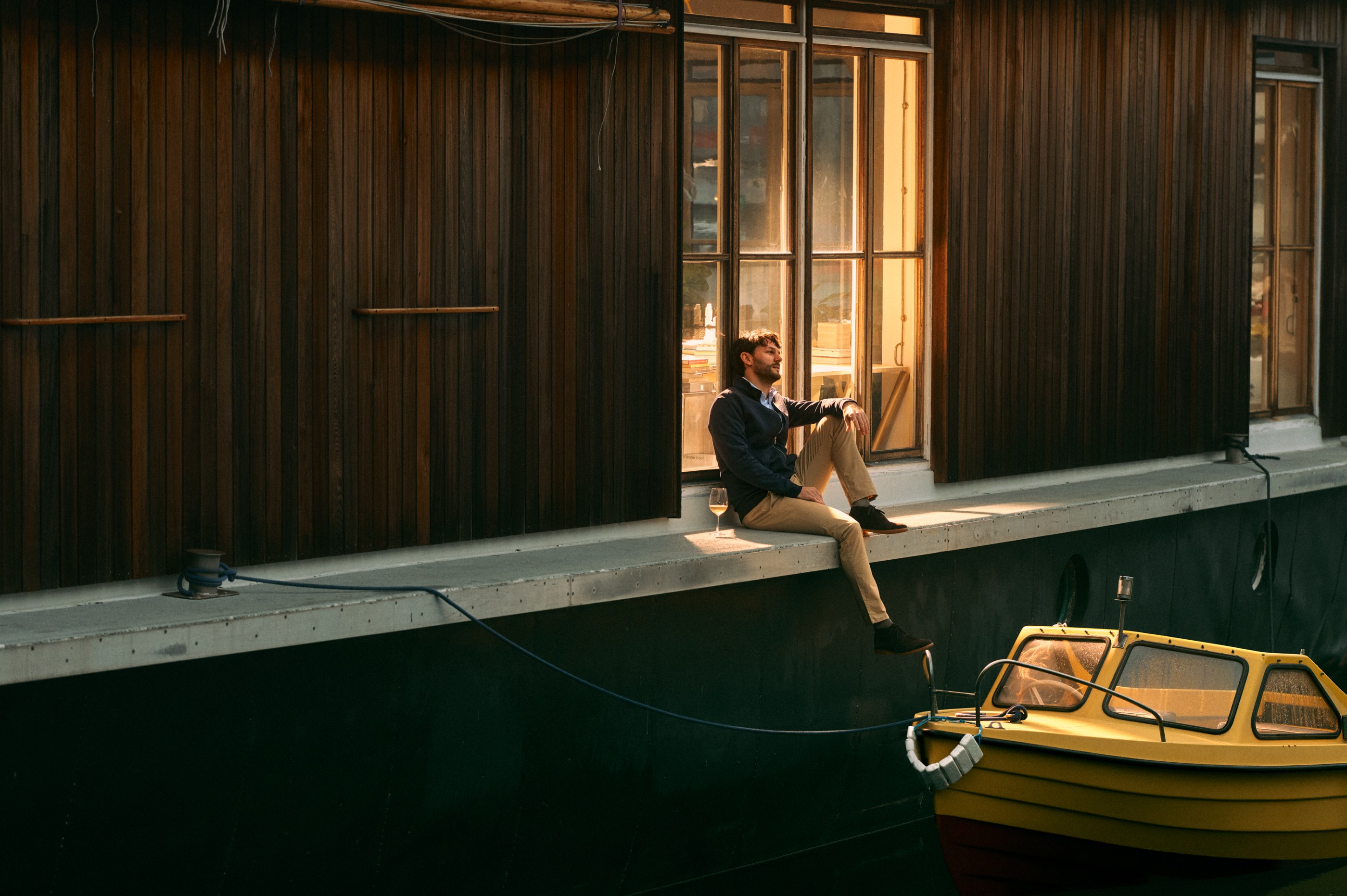 Man sitting on a dock with a glass of wine by a yellow boat at golden hour.