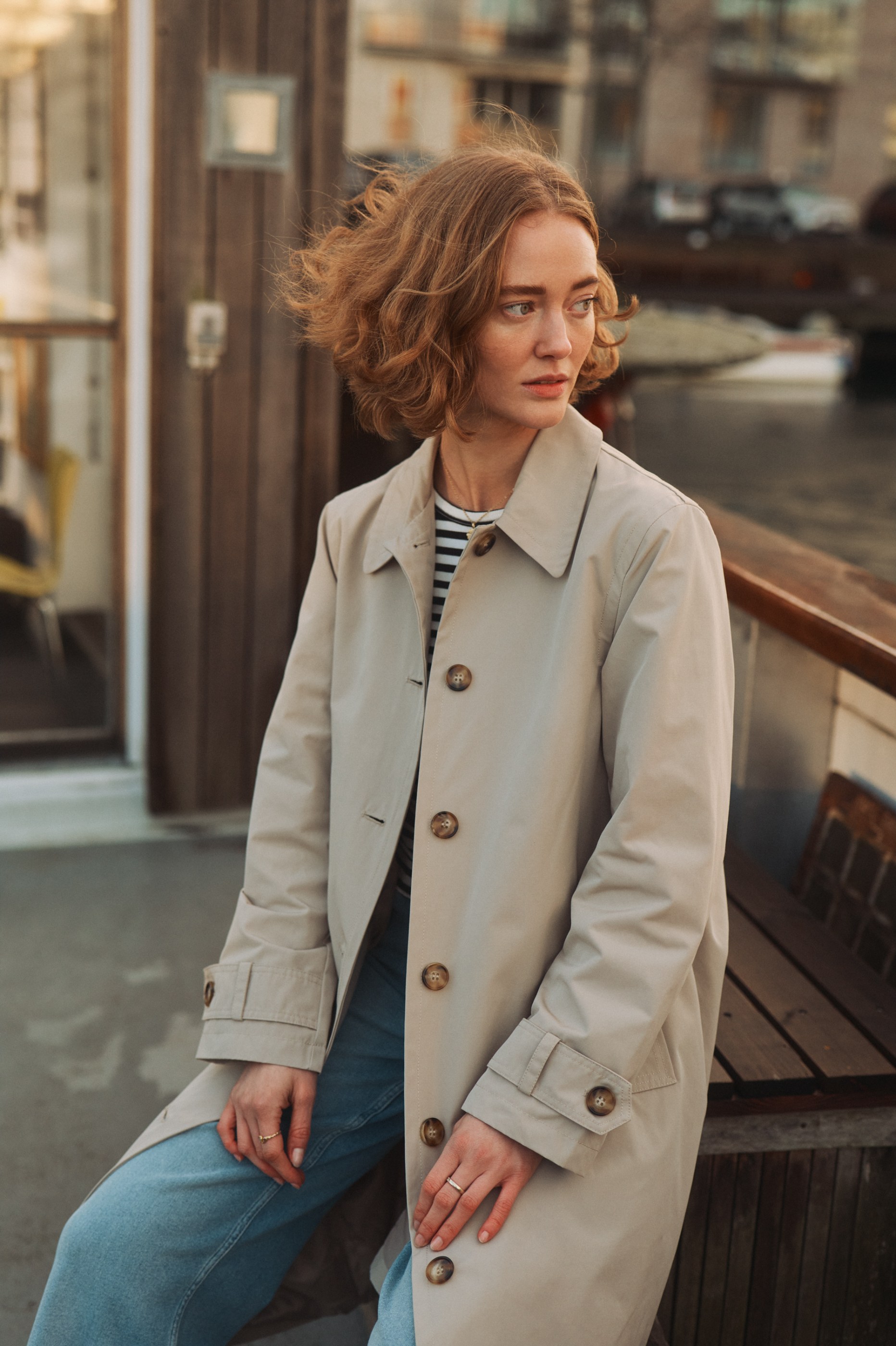 Pensive woman with wavy red hair in a beige trench coat sits on a wooden deck.