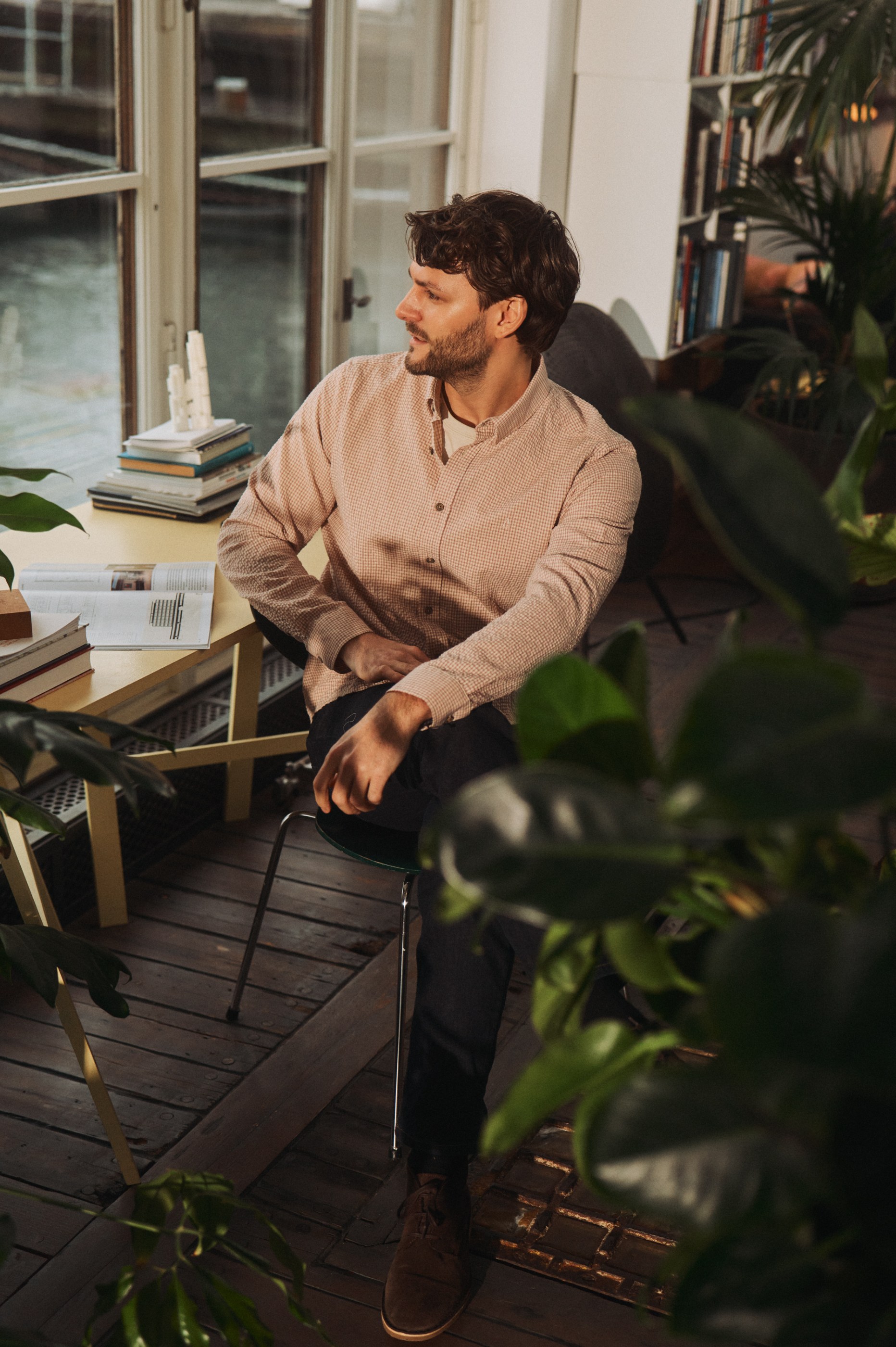 Thoughtful man in a pink checkered shirt sitting by a window in a plant-filled room.