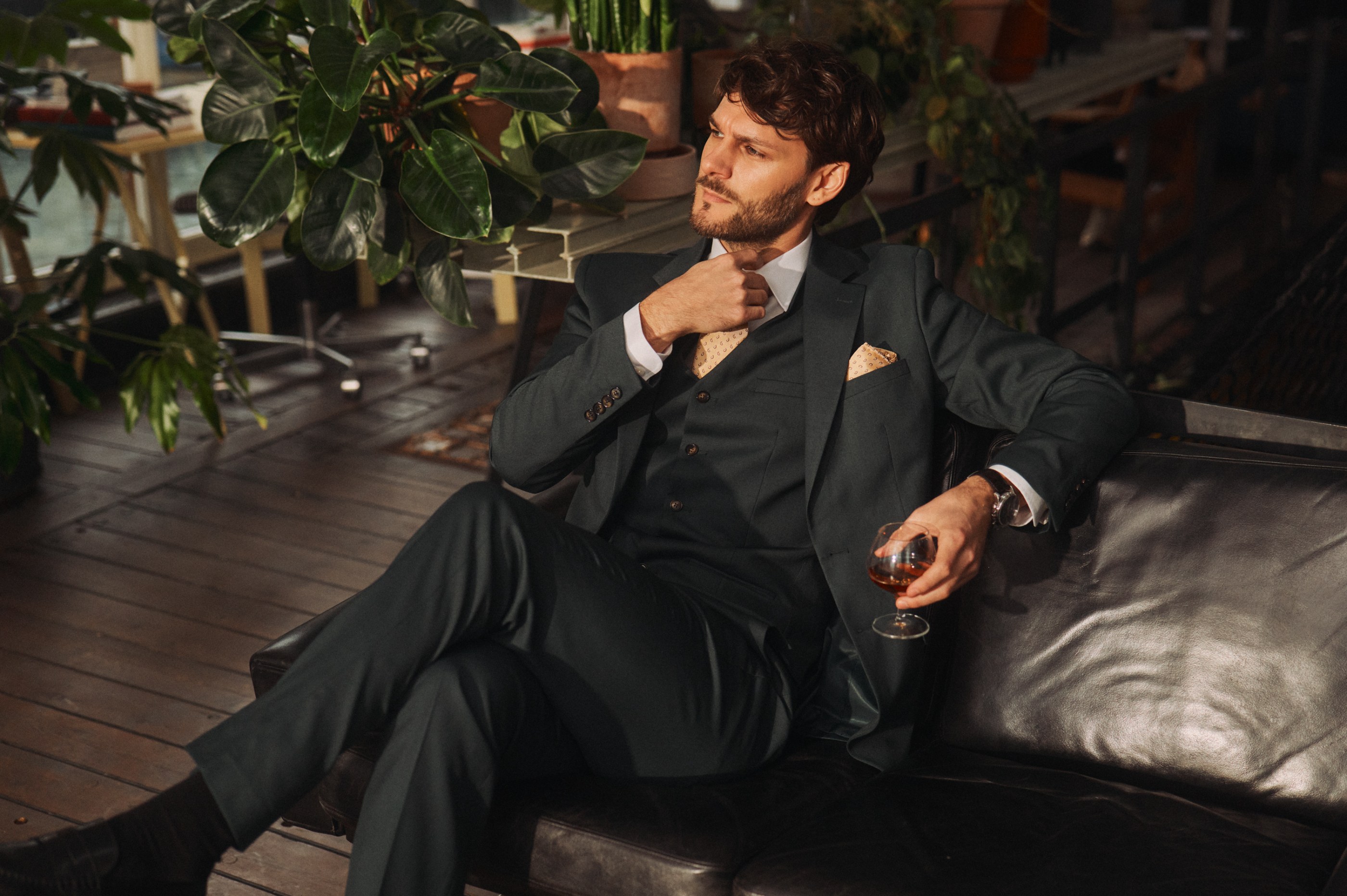 Man in a dark suit sits on a leather couch, holding a drink, surrounded by green plants.