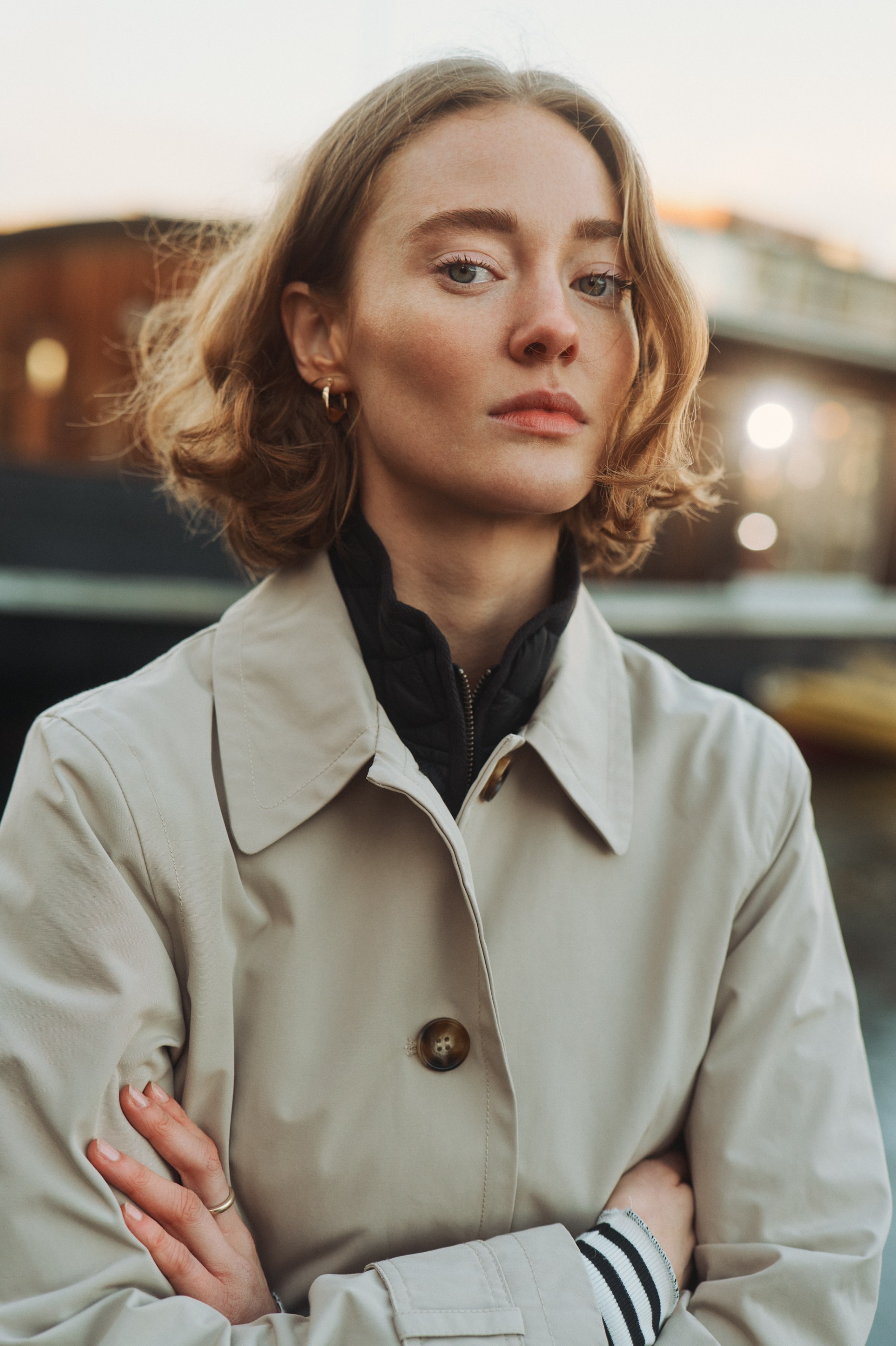 Blonde woman in beige trench coat, arms crossed, looking directly at the camera.