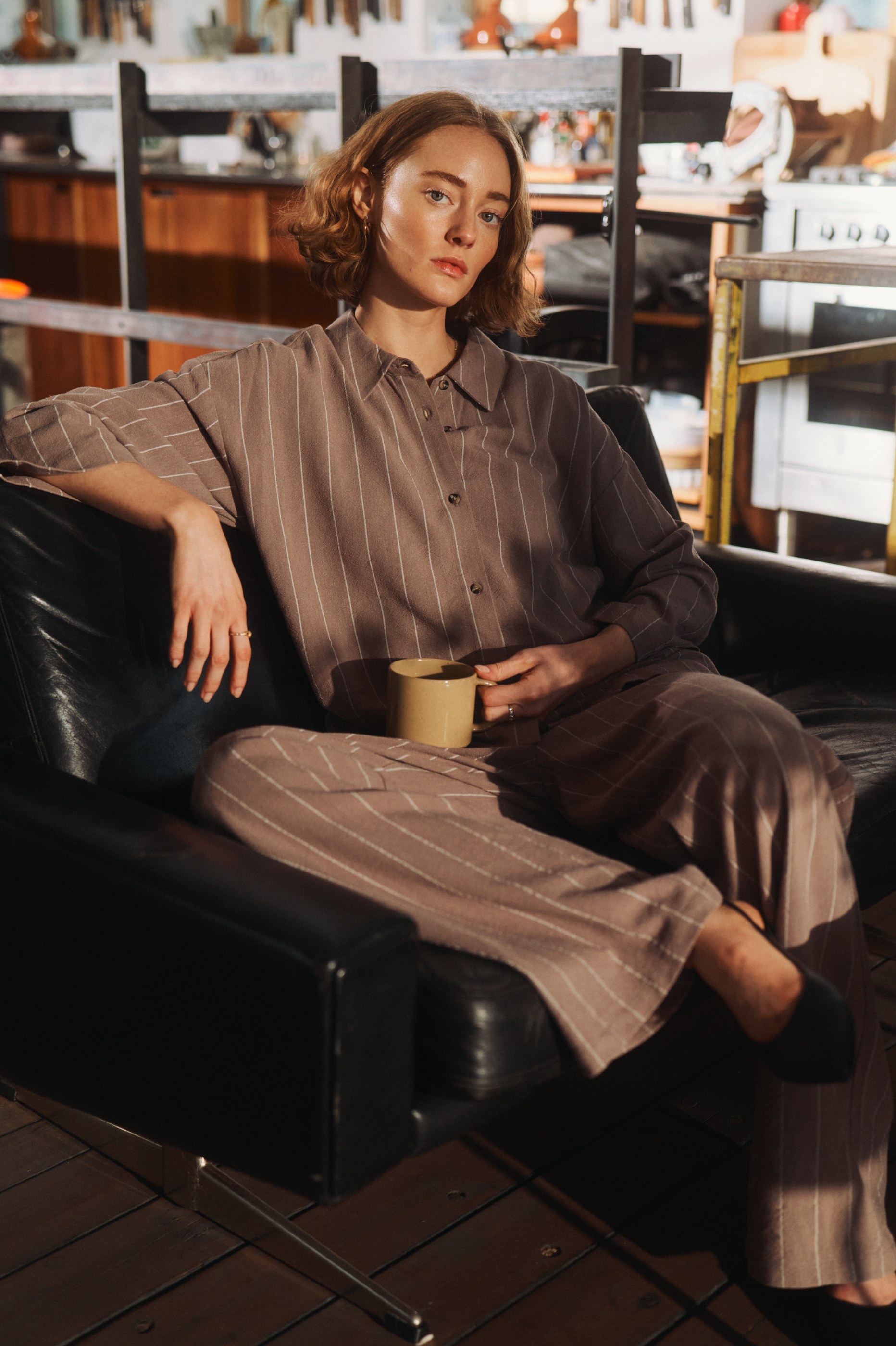 Woman in striped brown outfit sits on black leather couch, holding a mug.