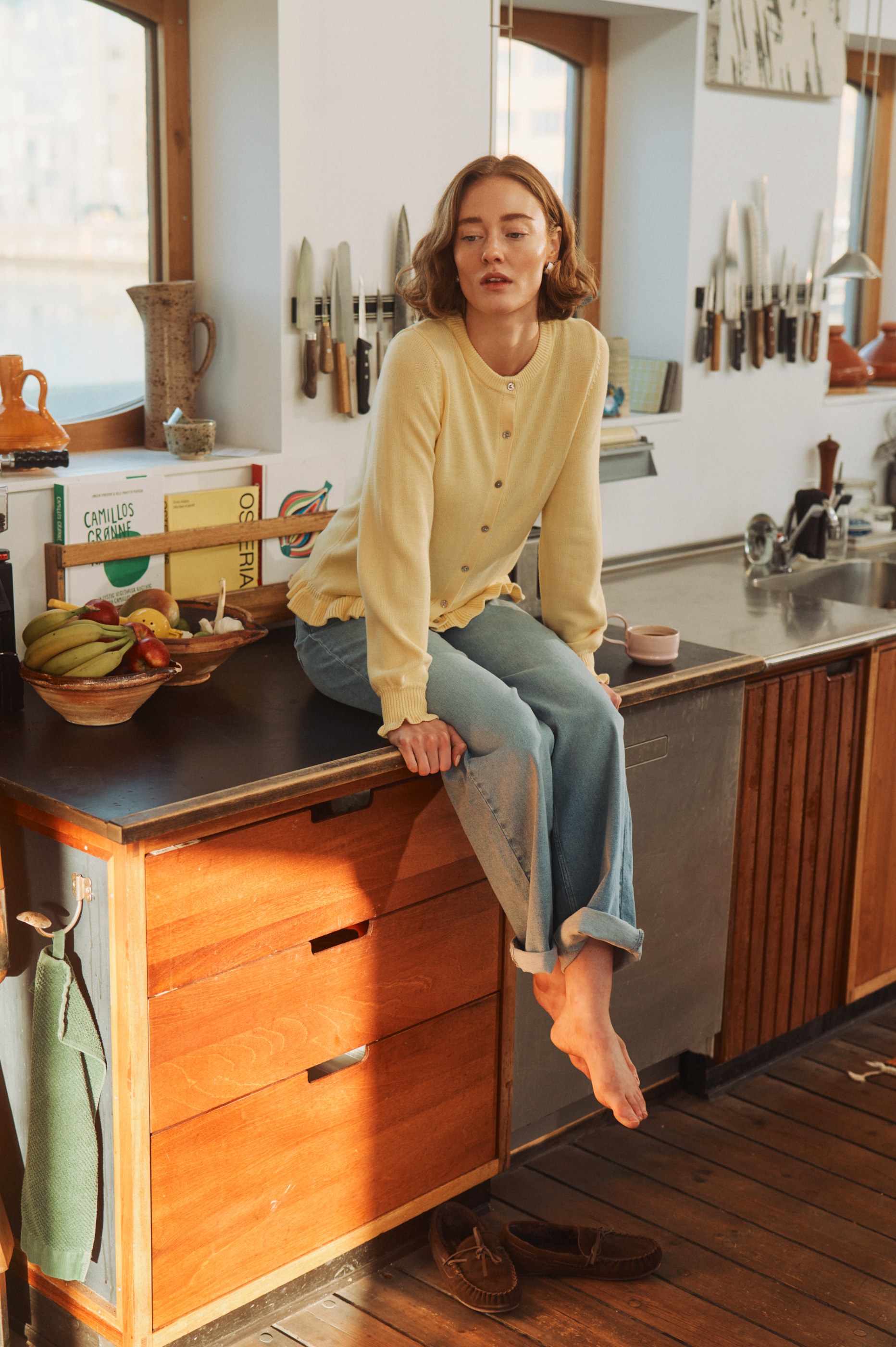 Barefoot woman in yellow cardigan and light blue jeans sits on a sunlit kitchen counter.