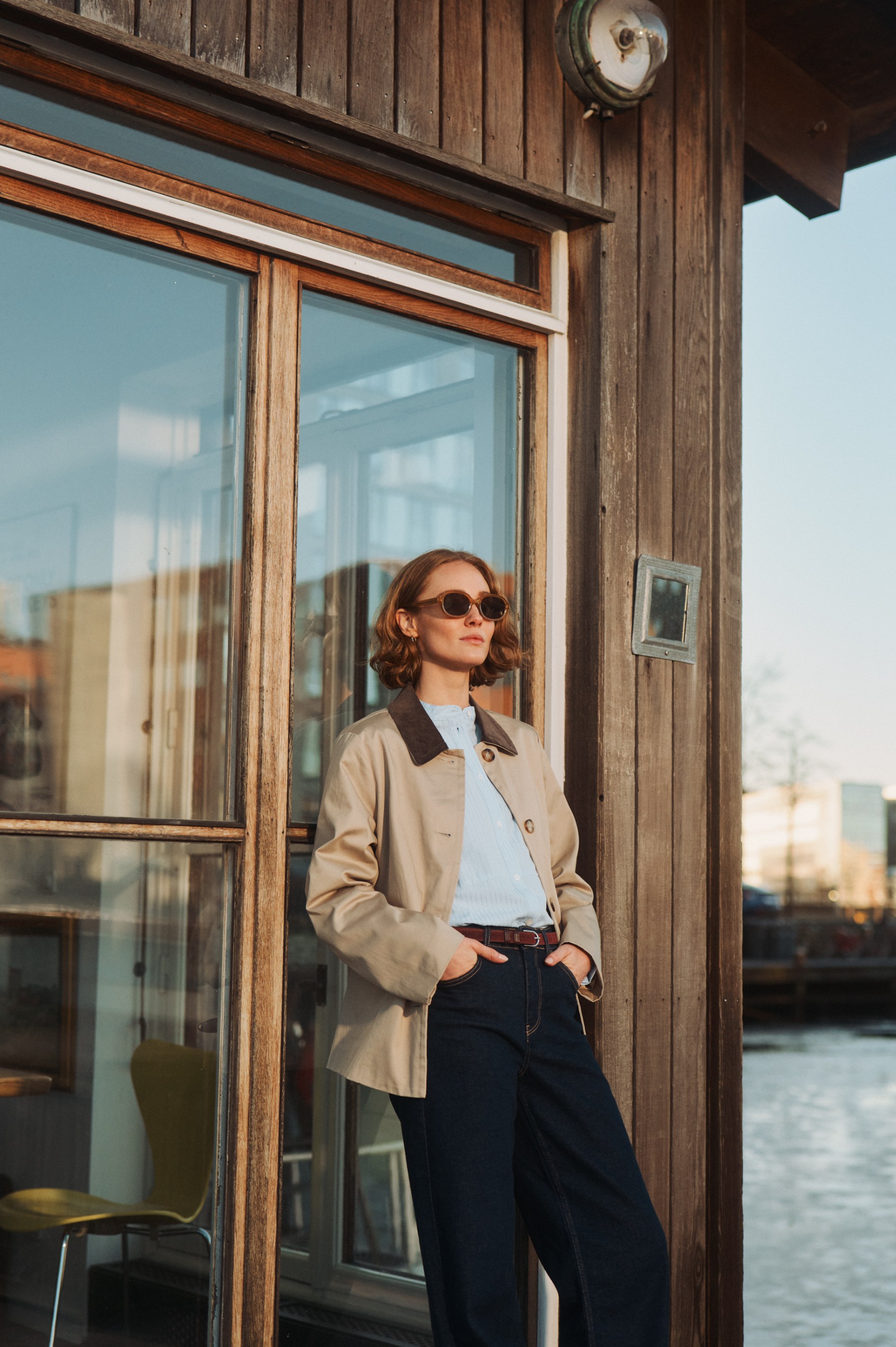 Stylish woman in sunglasses and a beige jacket leaning on a wooden building by water at sunset.
