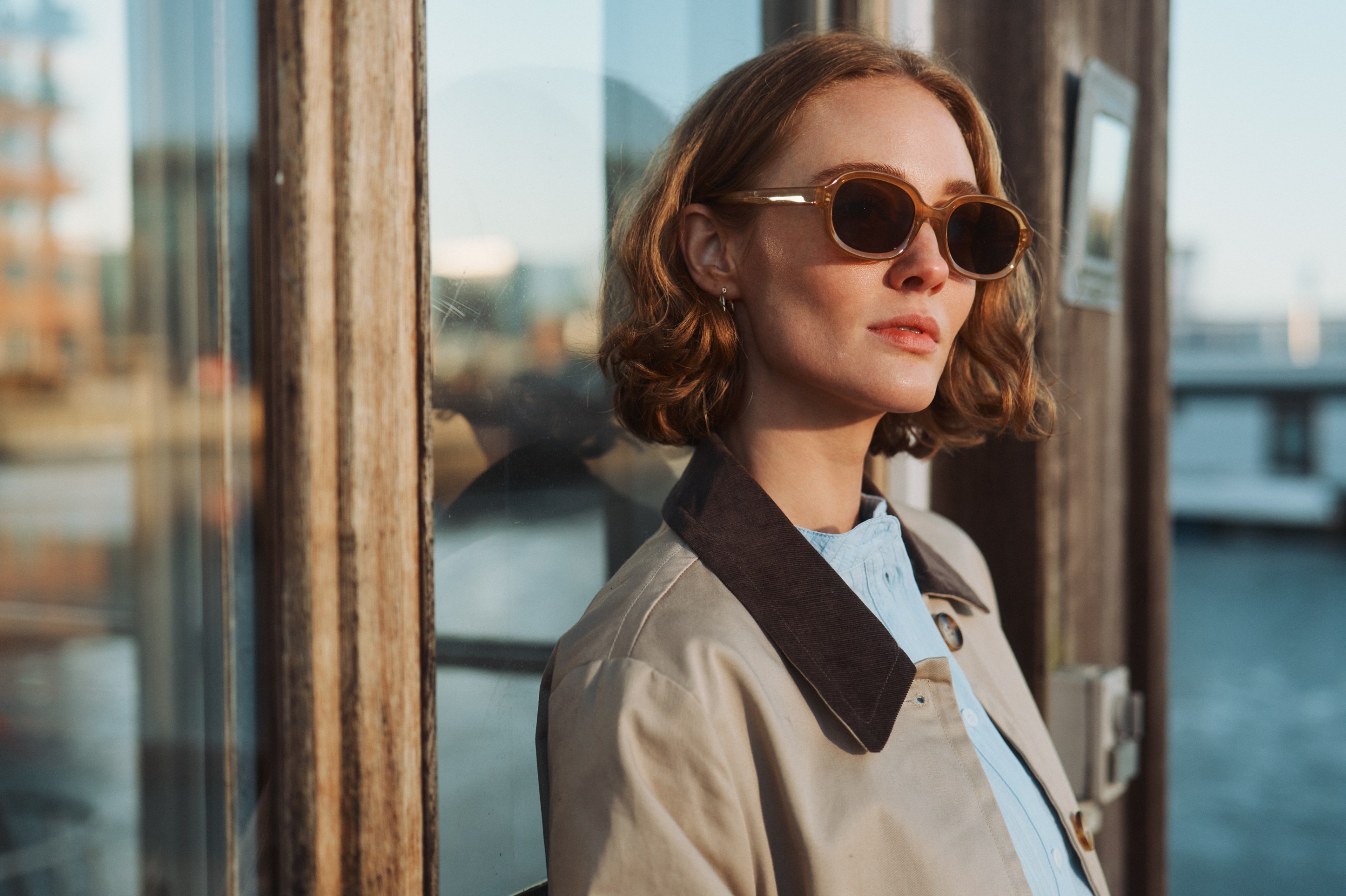 Woman in a trench coat and sunglasses, illuminated by golden light near a window