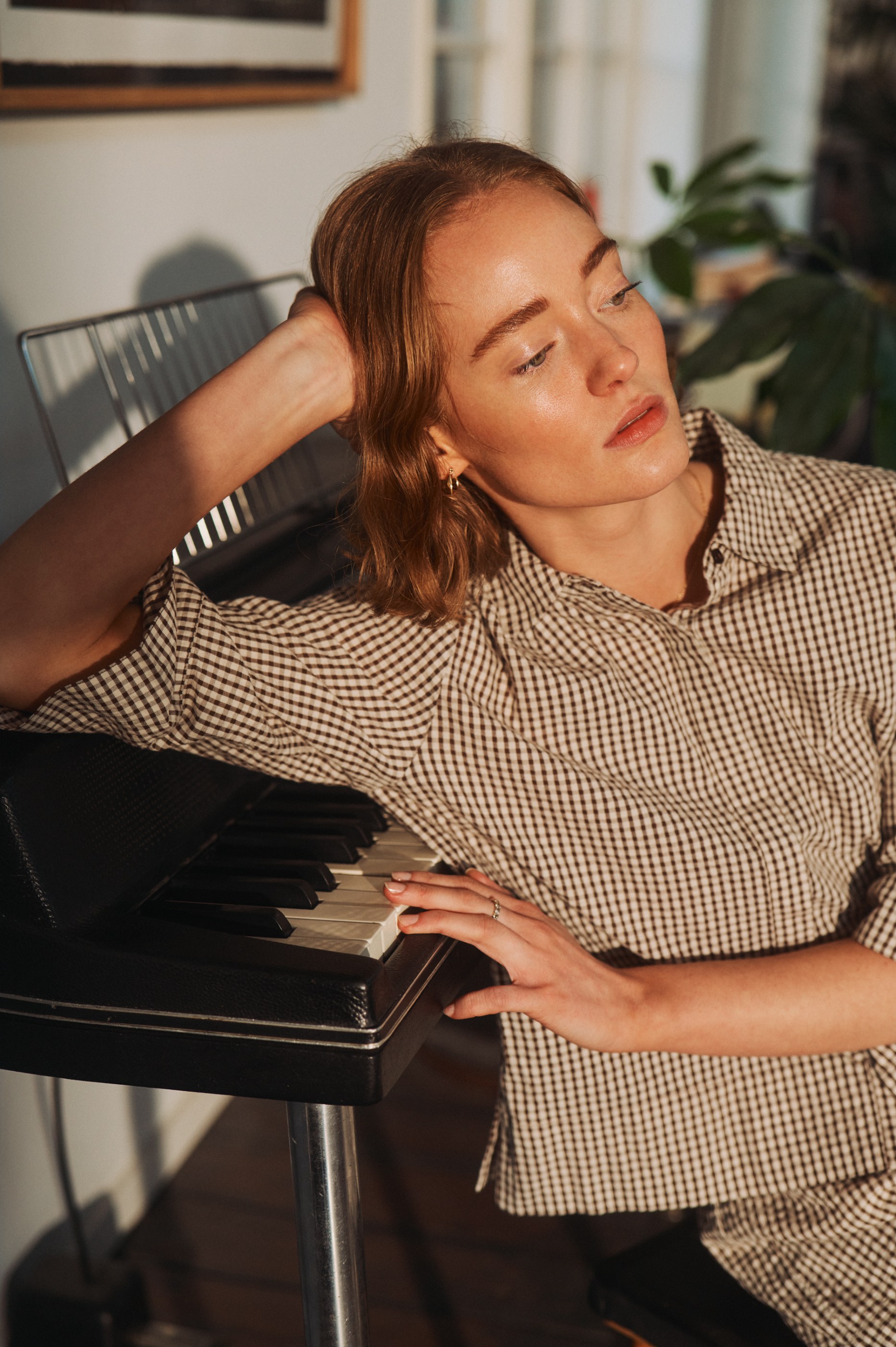 Woman in a plaid shirt with hand on piano keys, bathed in warm sunlight.