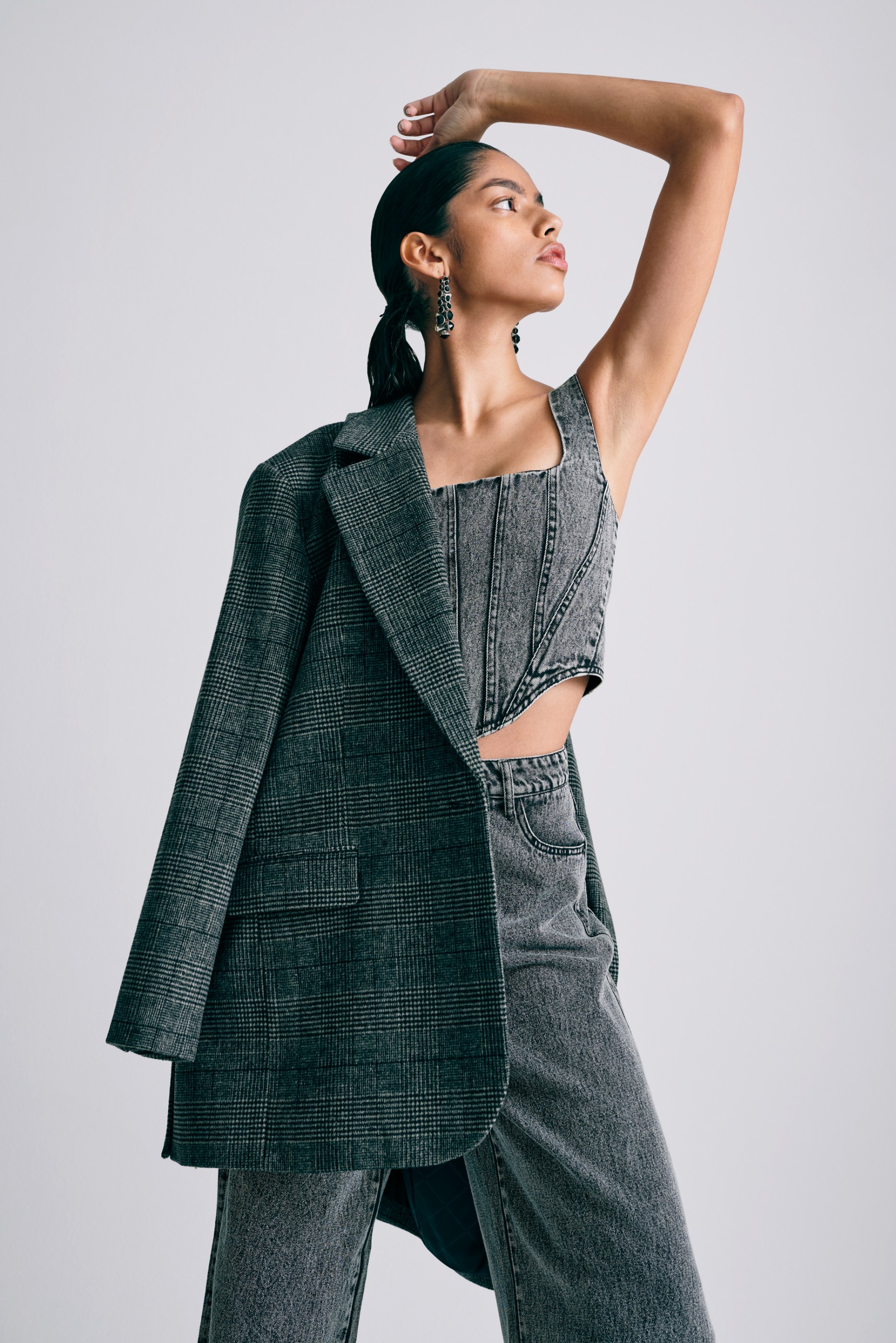 Woman posing in grey plaid blazer draped over shoulder, denim corset top, and jeans.