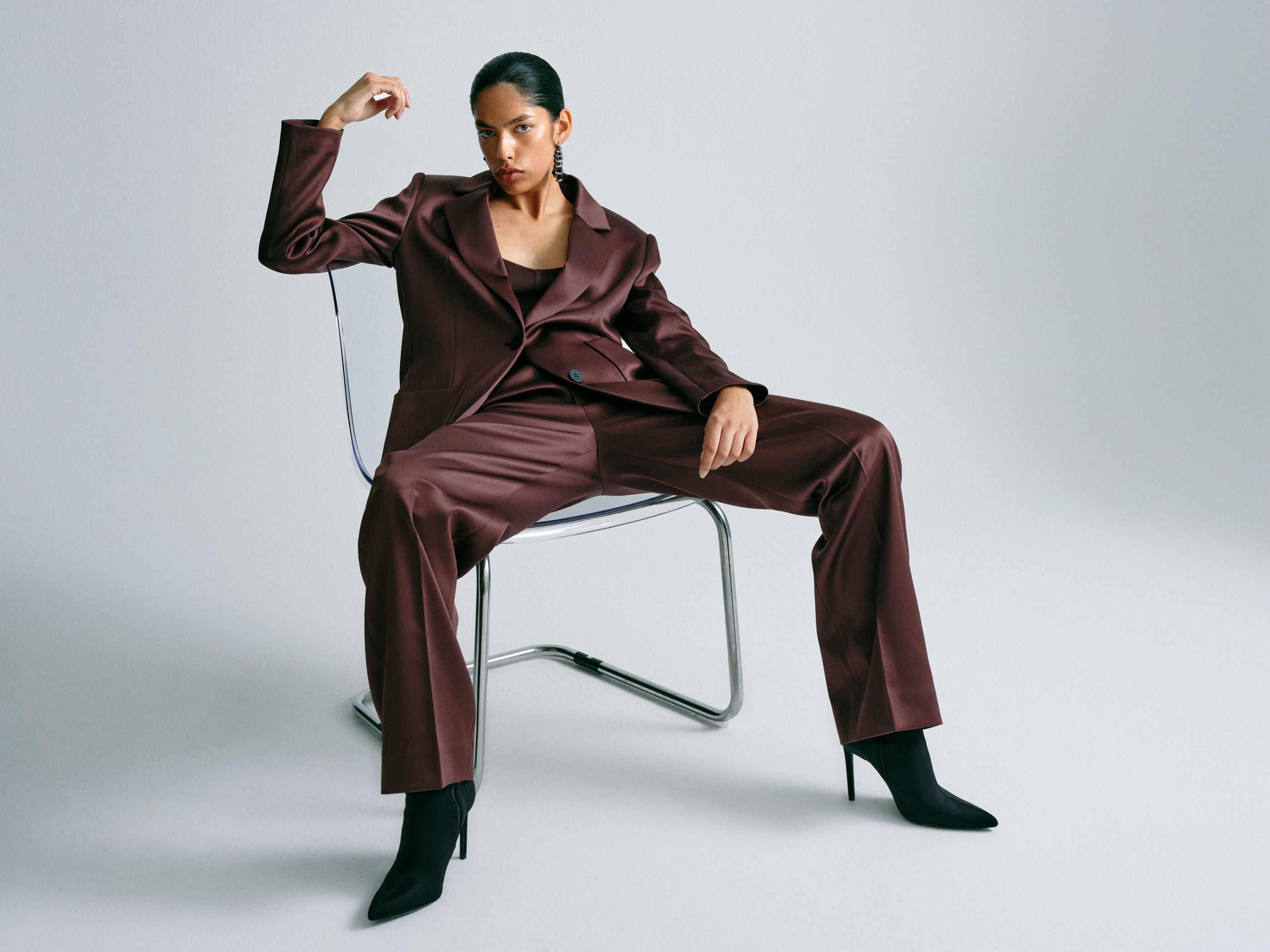 Confident woman in a dark brown satin suit and black boots sitting on a transparent chair.