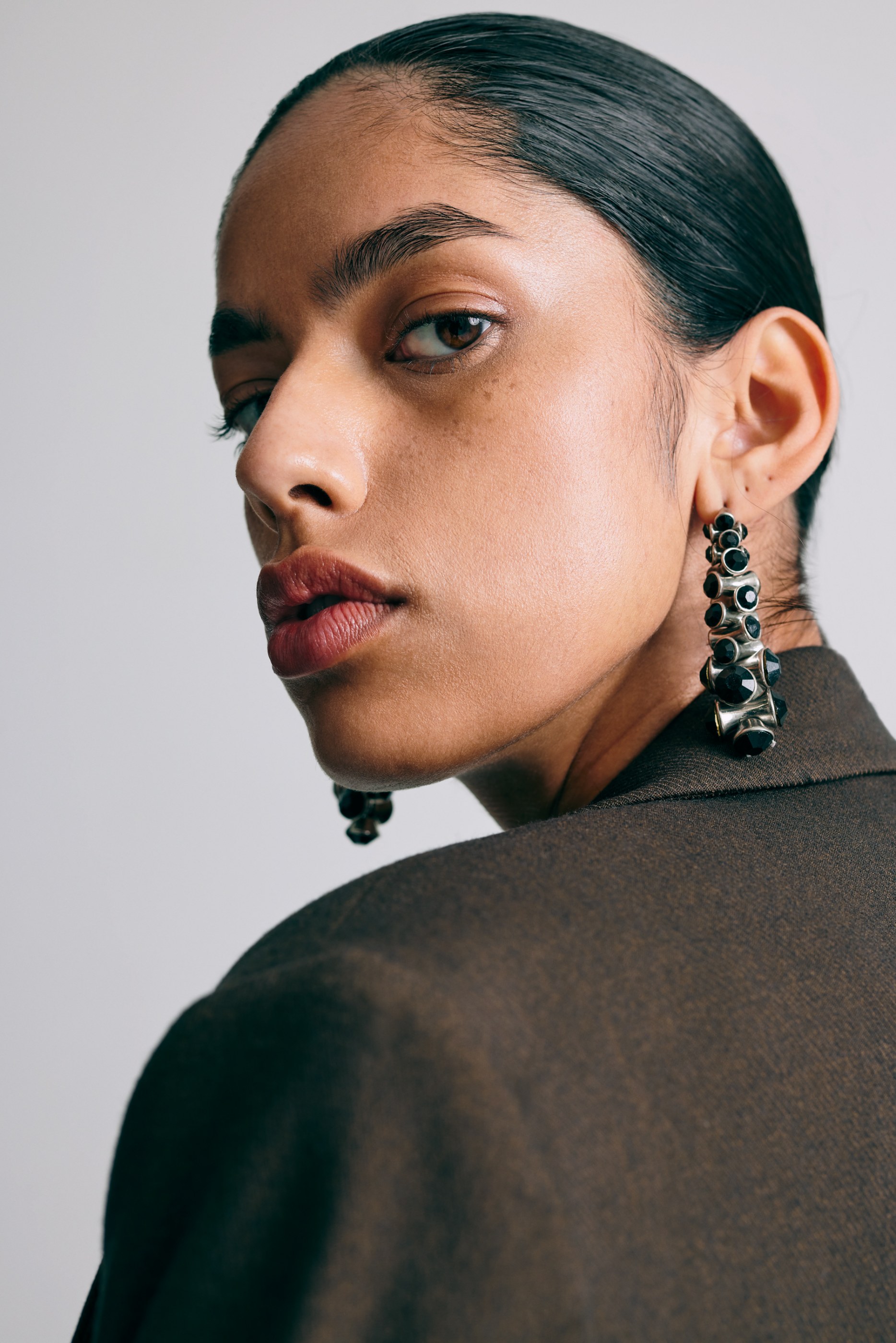 Woman in a brown blazer, with slicked dark hair and black drop earrings, looking back.