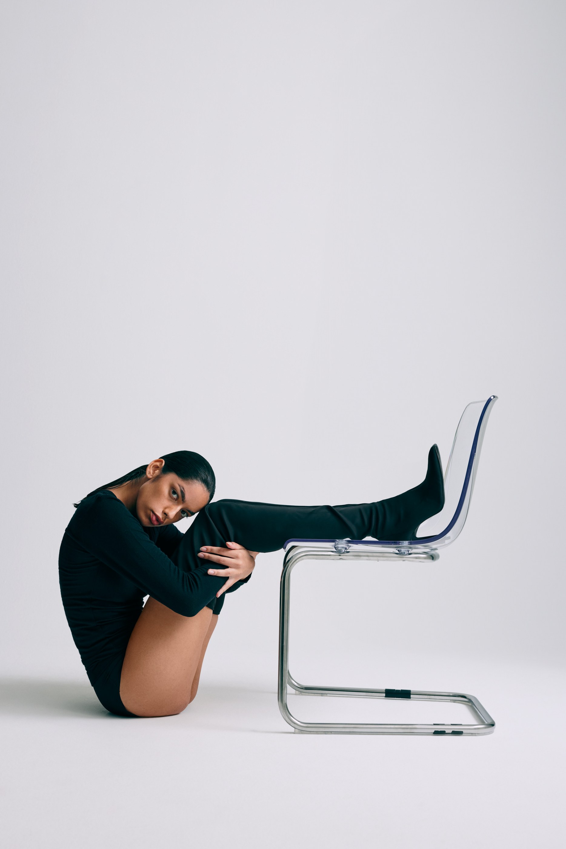 Woman in black bodysuit sitting on floor with leg on transparent chair in a white studio.
