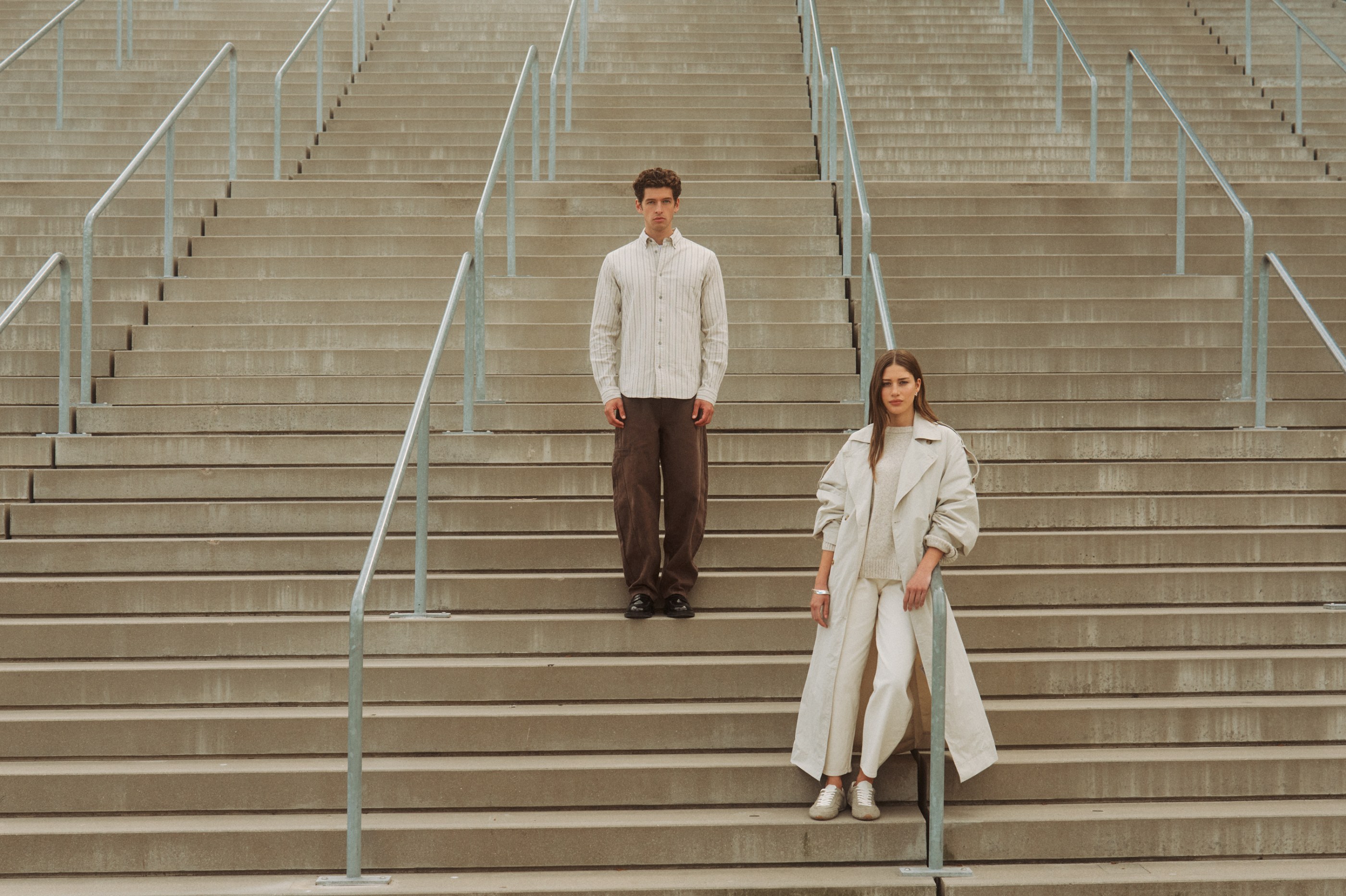 A man and a woman in neutral outfits stand on a wide concrete staircase with railings.
