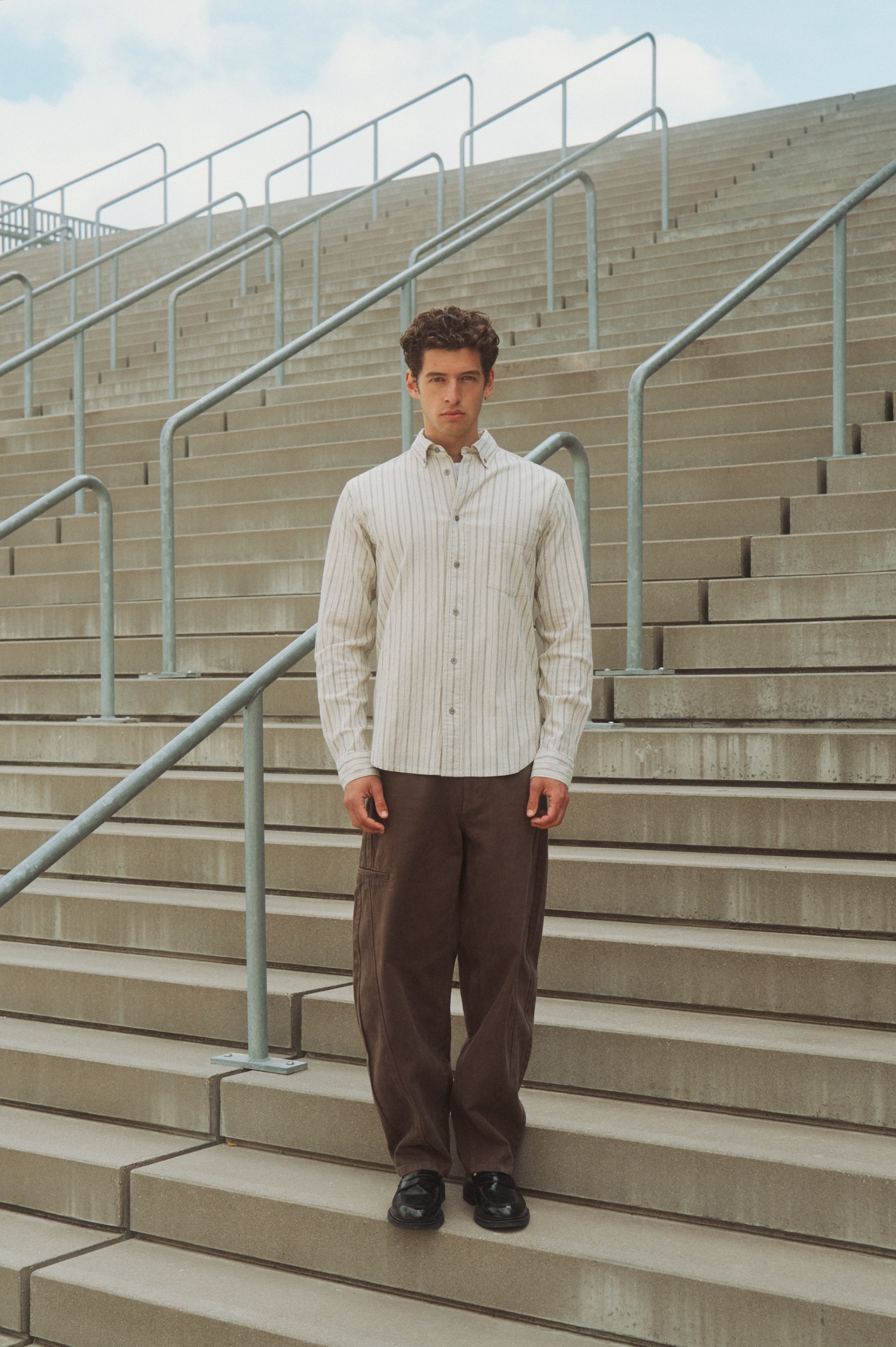 Man in a striped shirt, brown pants, and black shoes stands on concrete stairs with railings.