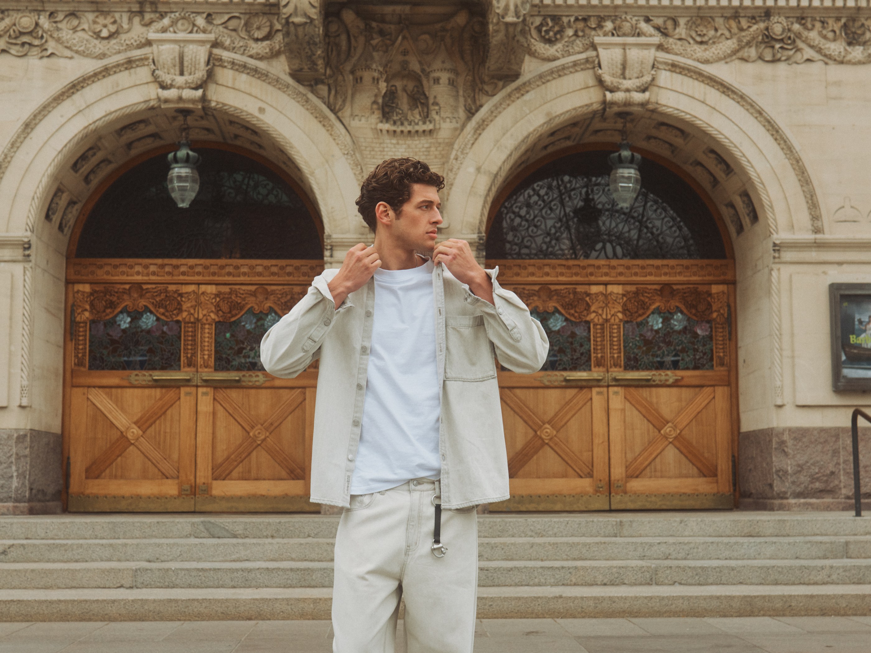 Man in light shirt jacket and pants adjusting collar in front of an ornate arched building entrance.
