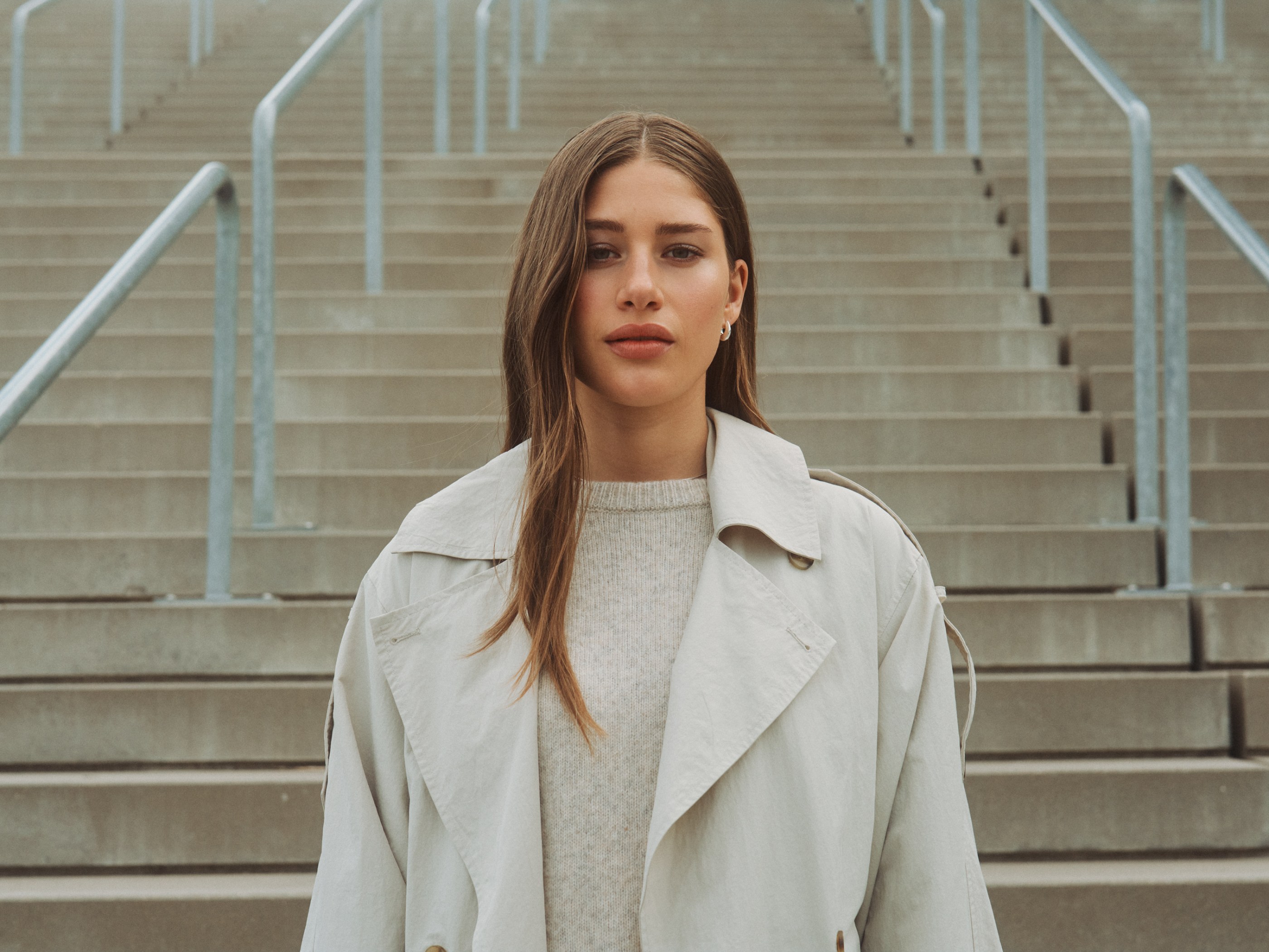 Young woman in a light trench coat standing on concrete stairs with railings, looking at the camera.