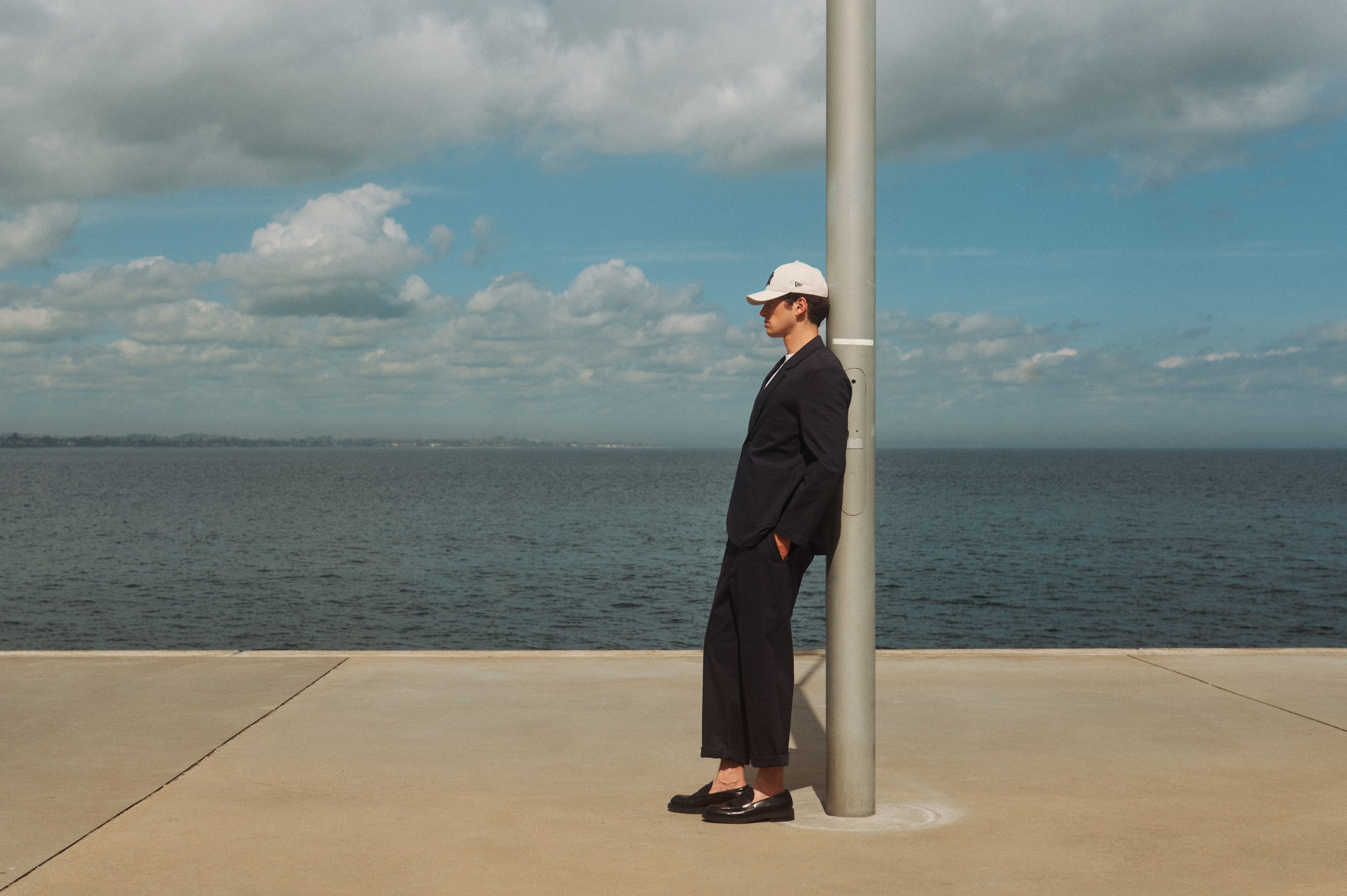 Man in a dark suit and white baseball cap leaning on a pole by the ocean.