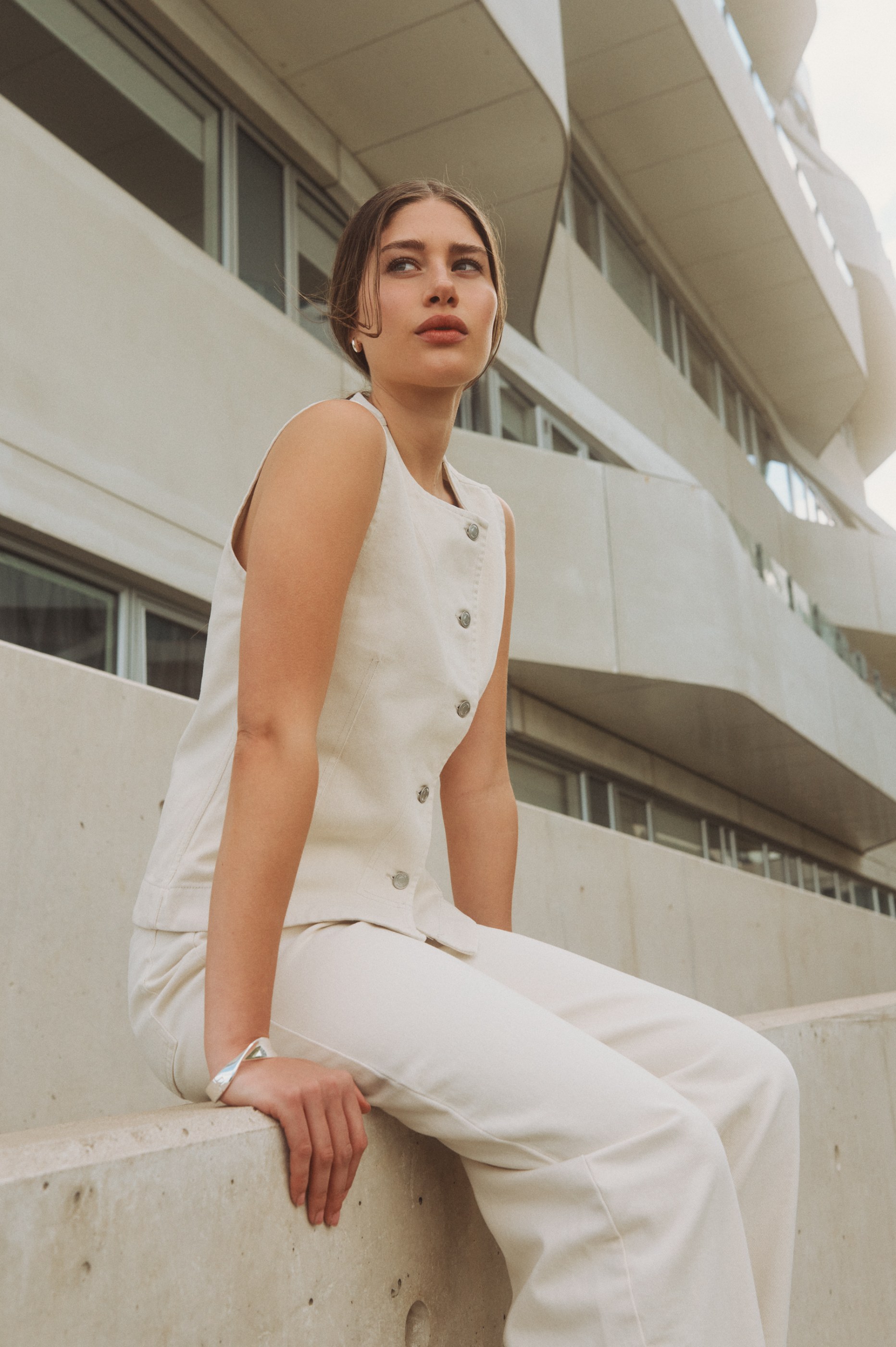 Elegant woman in cream outfit sitting on concrete, looking up at a modern building.