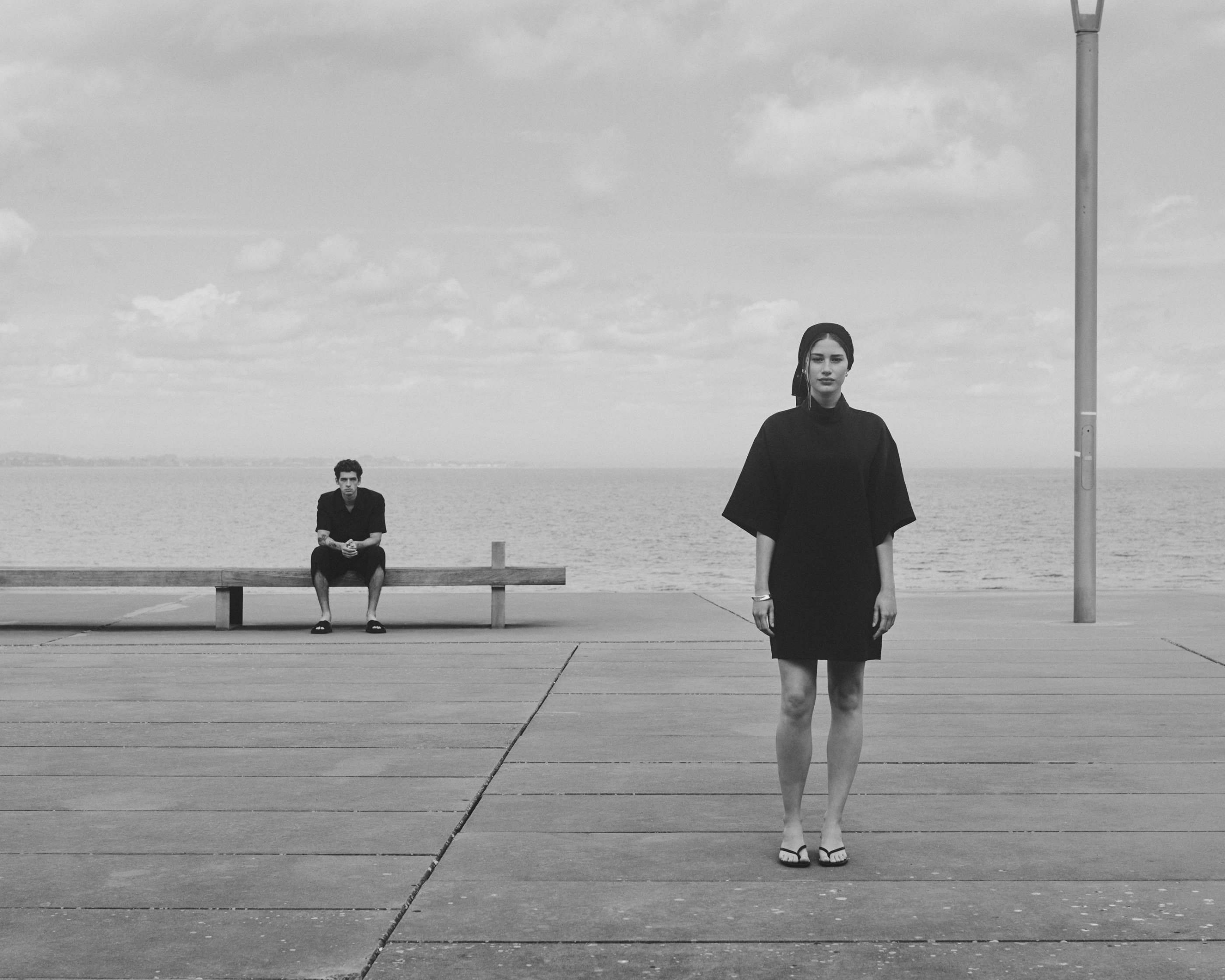 Monochrome: man on bench, woman standing on pier by the sea.