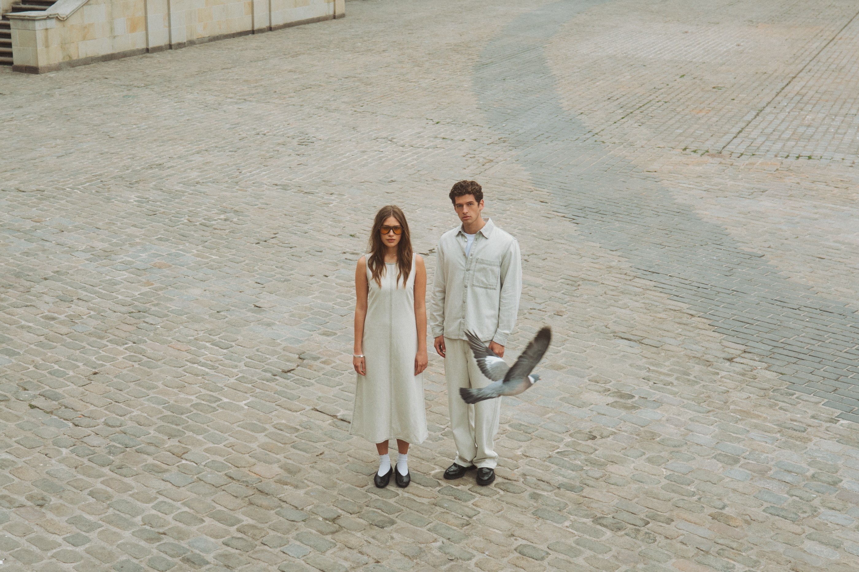 Man and woman in light clothing stand on cobblestones as a pigeon flies by.