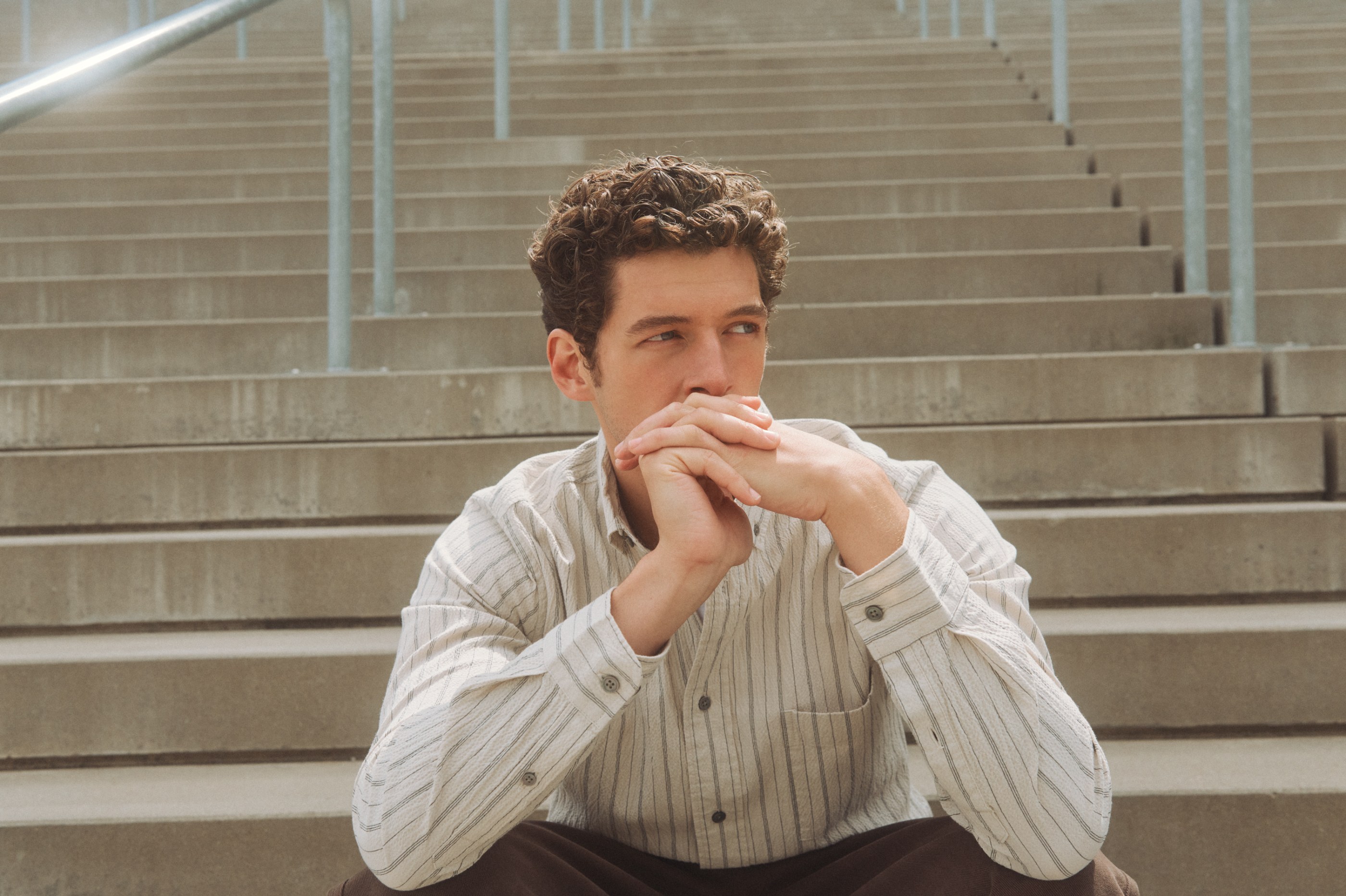Young man with curly hair in a striped shirt sitting pensively on concrete steps.