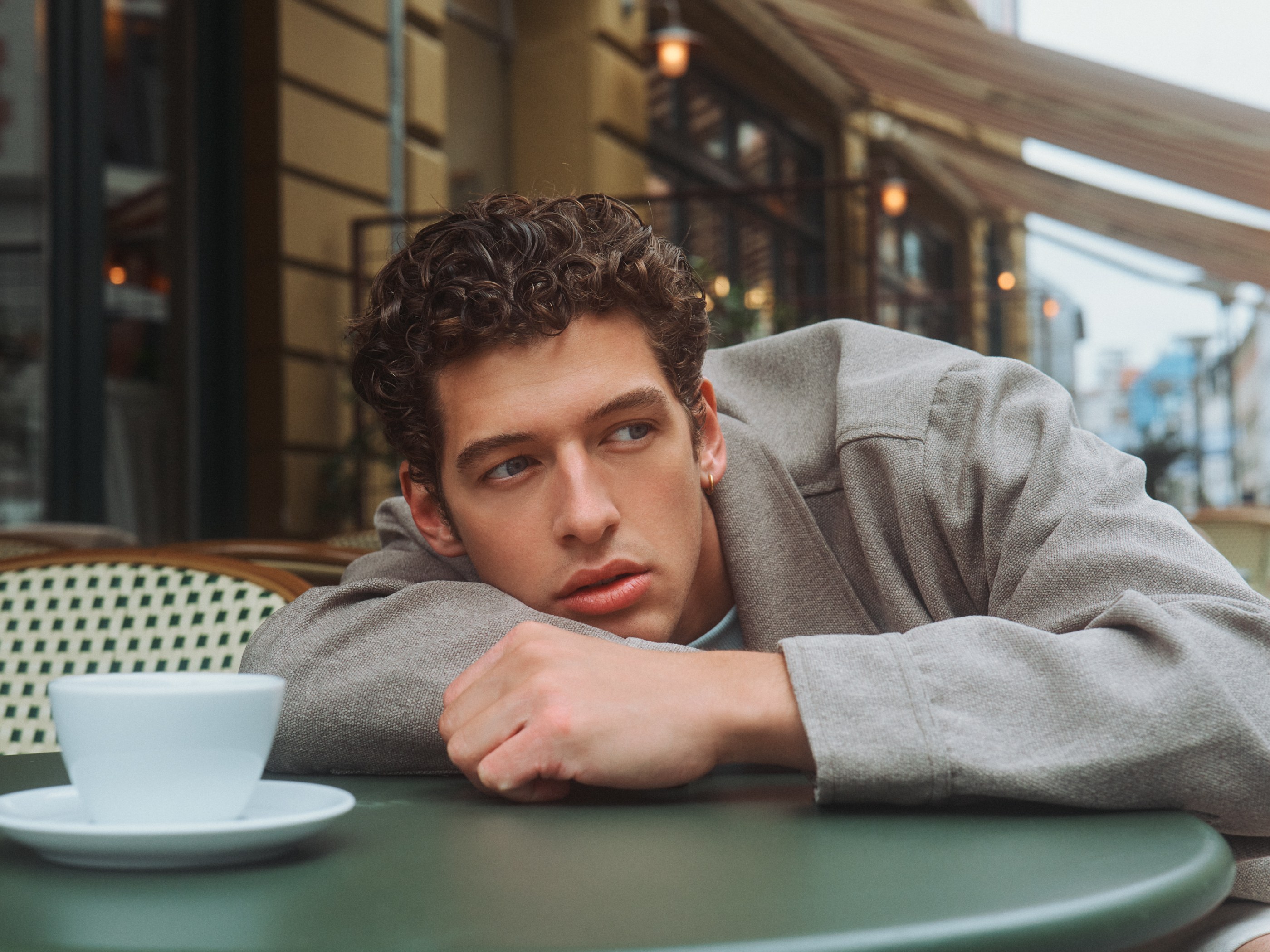Thoughtful young man in a gray jacket leaning on a cafe table with a white cup.