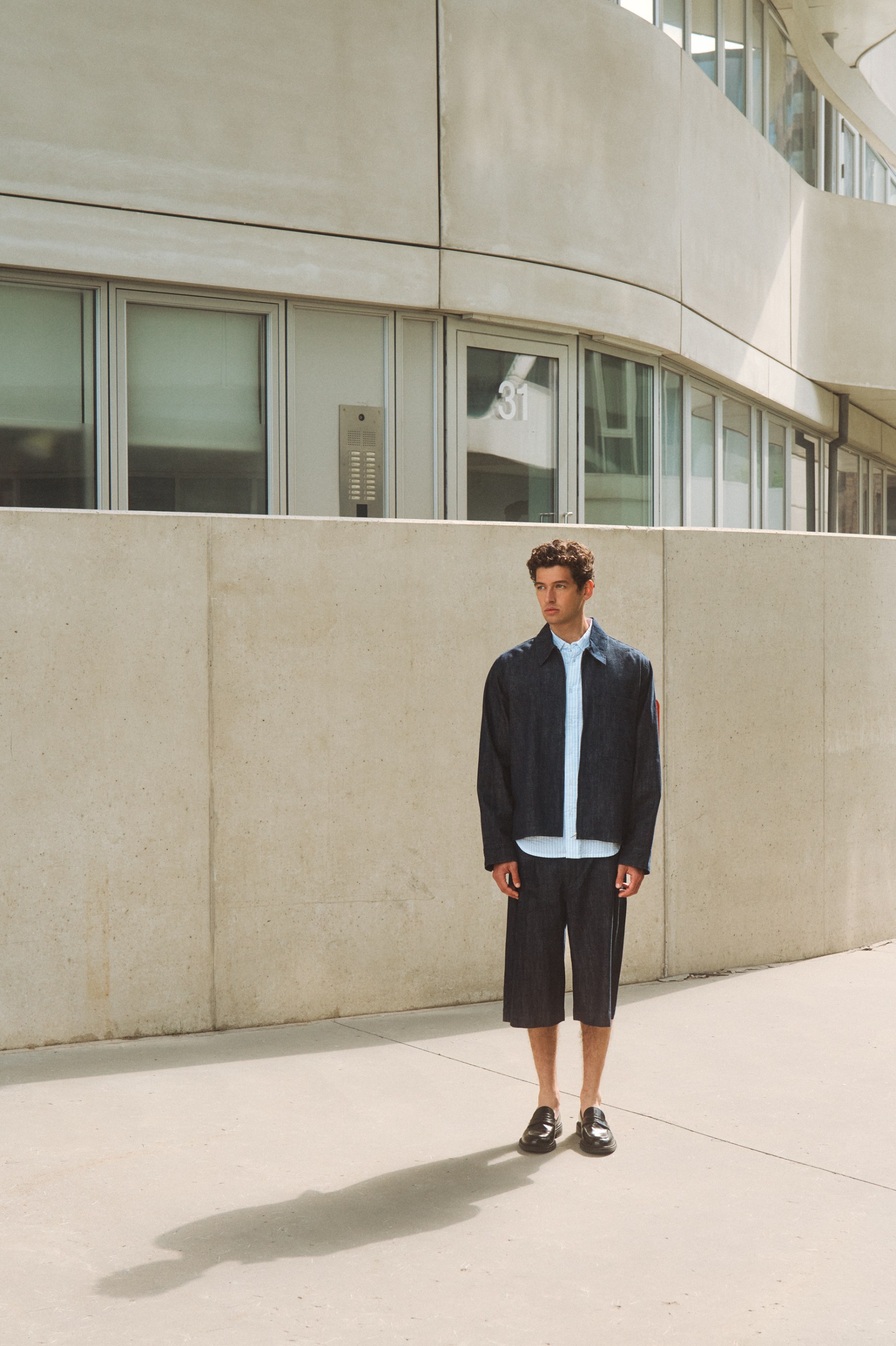Man in dark denim jacket, shorts, light shirt, and loafers by a modern building.