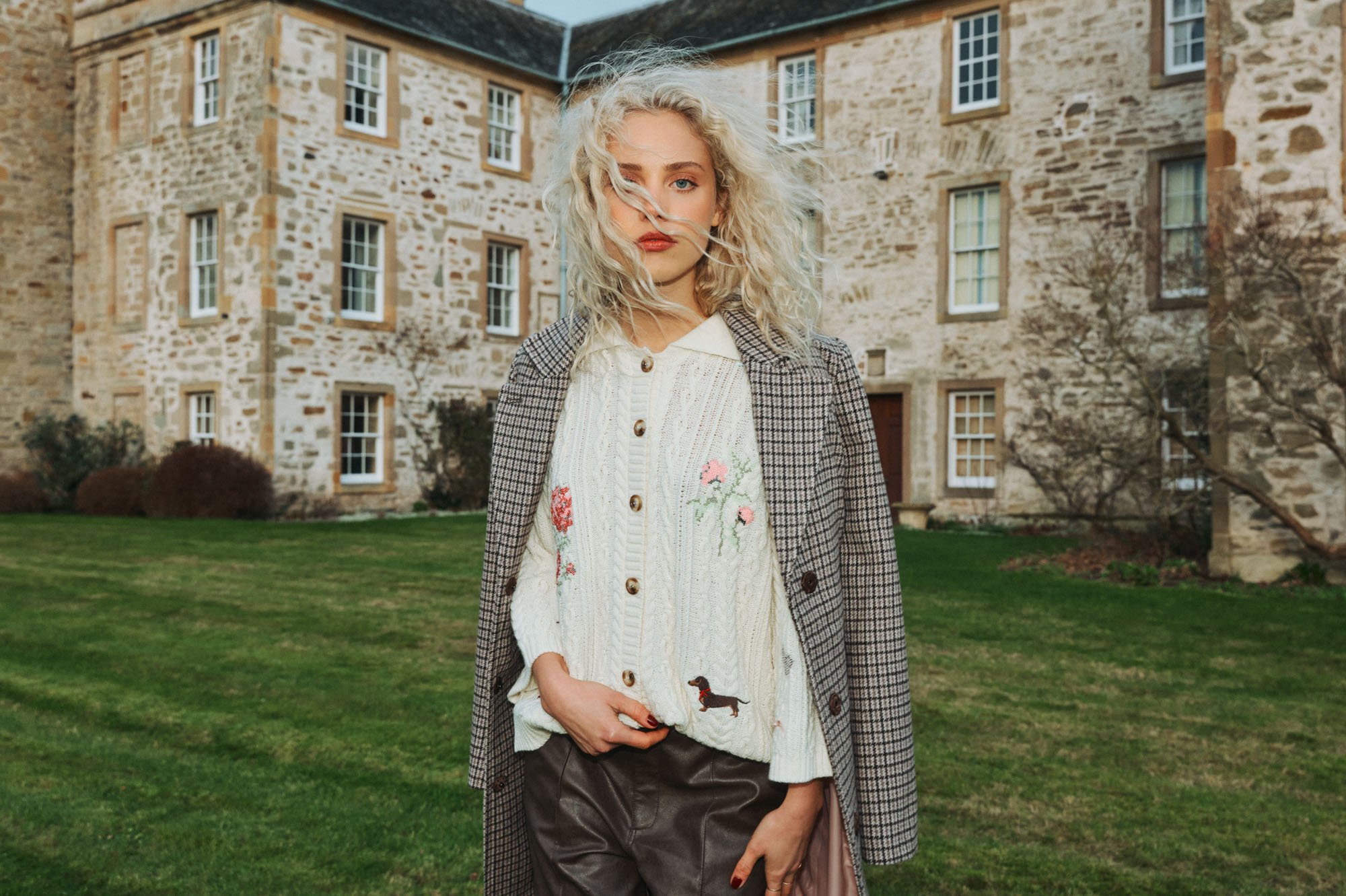 Blonde woman in plaid coat and embroidered cardigan on a lawn near a historic stone building.