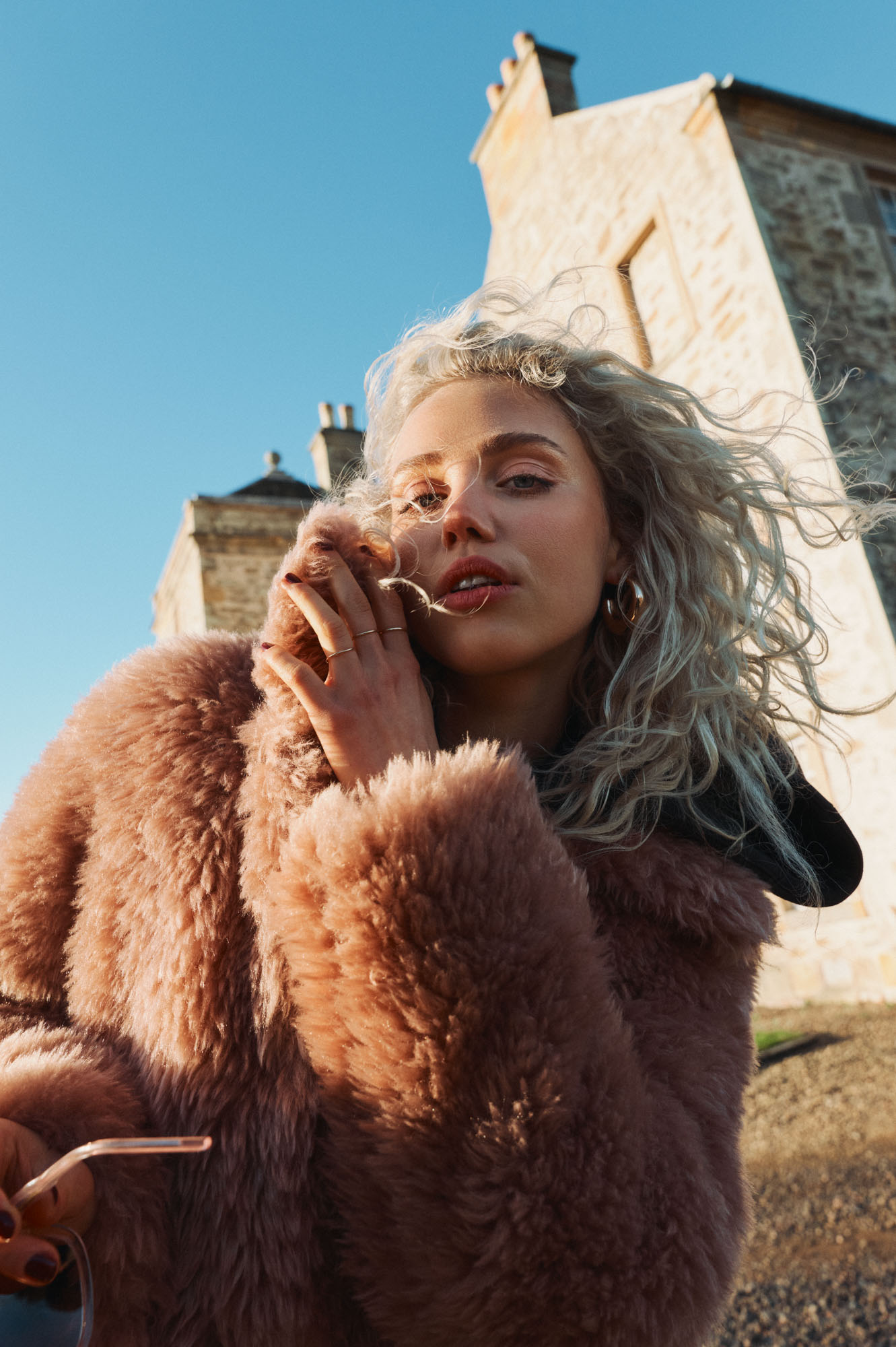 Windswept blonde woman in a pink fluffy coat looks at camera, with an old stone building.
