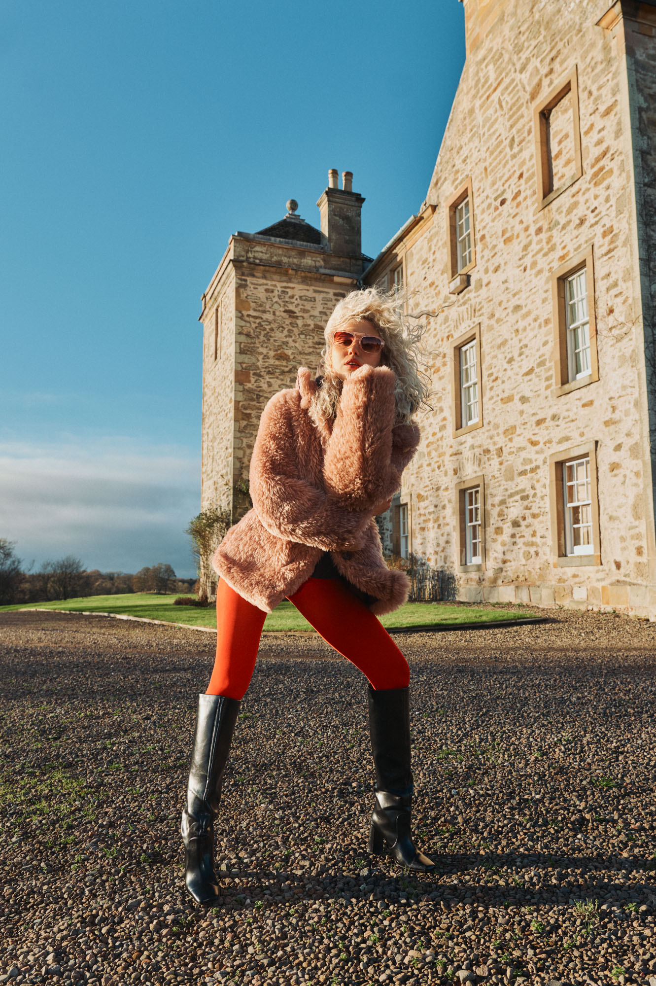 Confident woman in pink fur coat, red leggings, and black boots by a stone building.