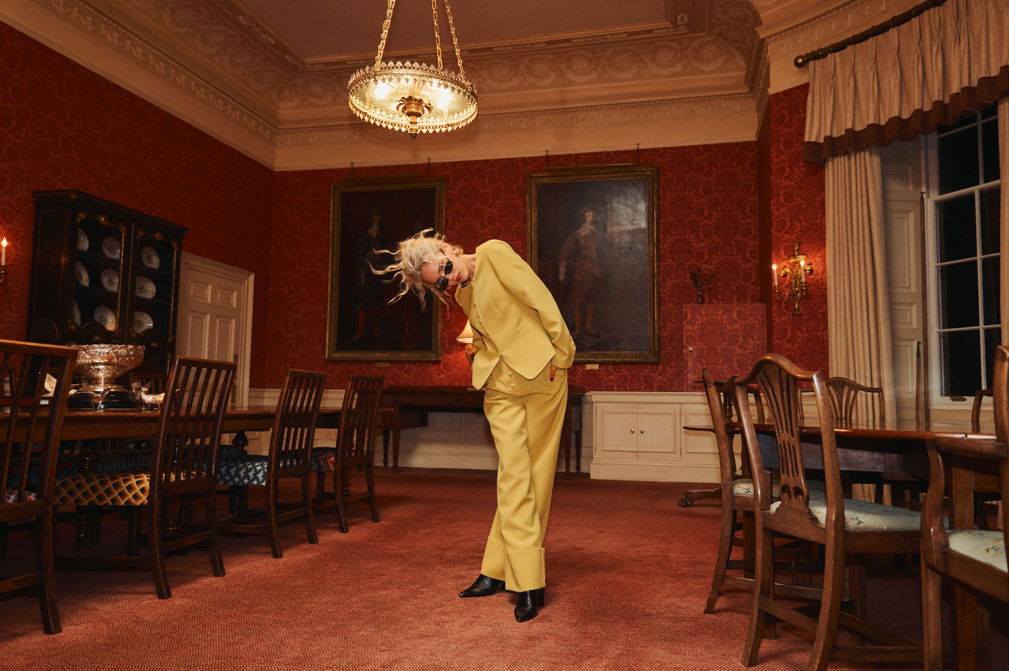 Person in yellow suit and sunglasses dramatically posing in an ornate red dining room.
