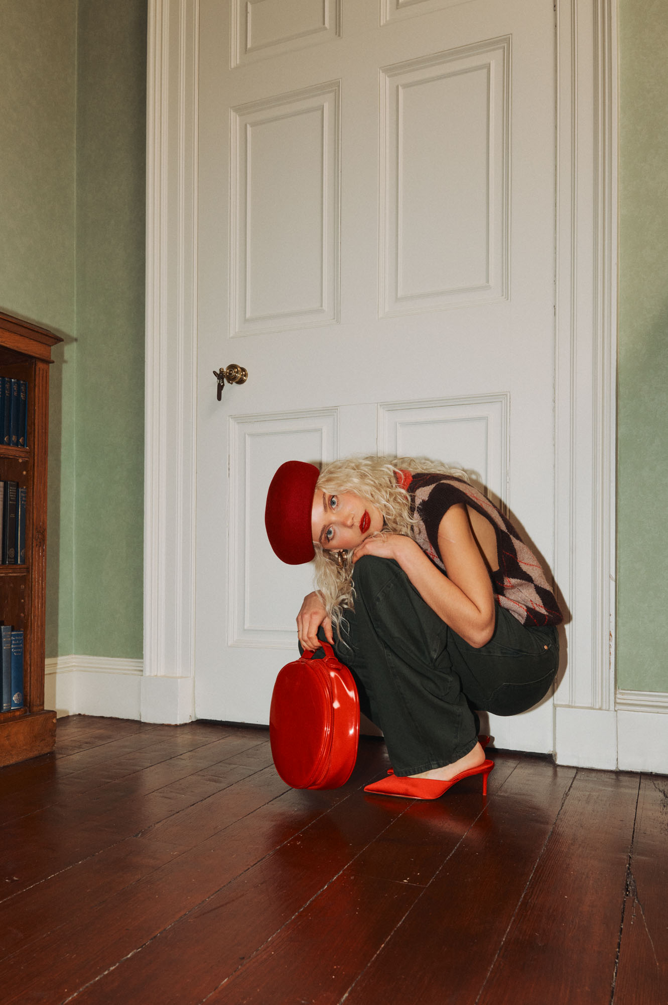 Blonde woman in red beret, argyle vest, and red heels crouching with a red bag.