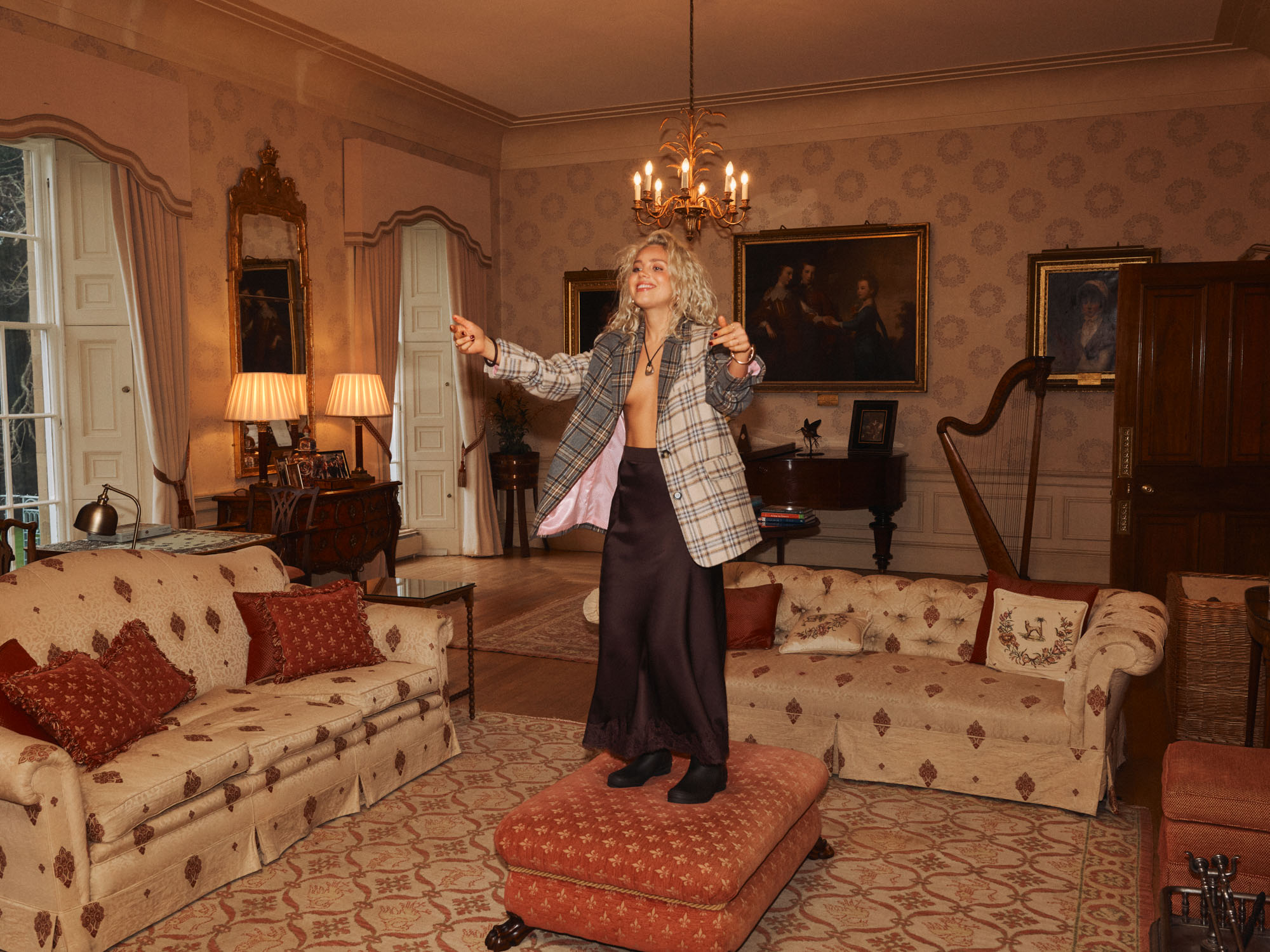 Smiling woman in plaid jacket and dark skirt standing on ottoman in ornate, warm-toned room.