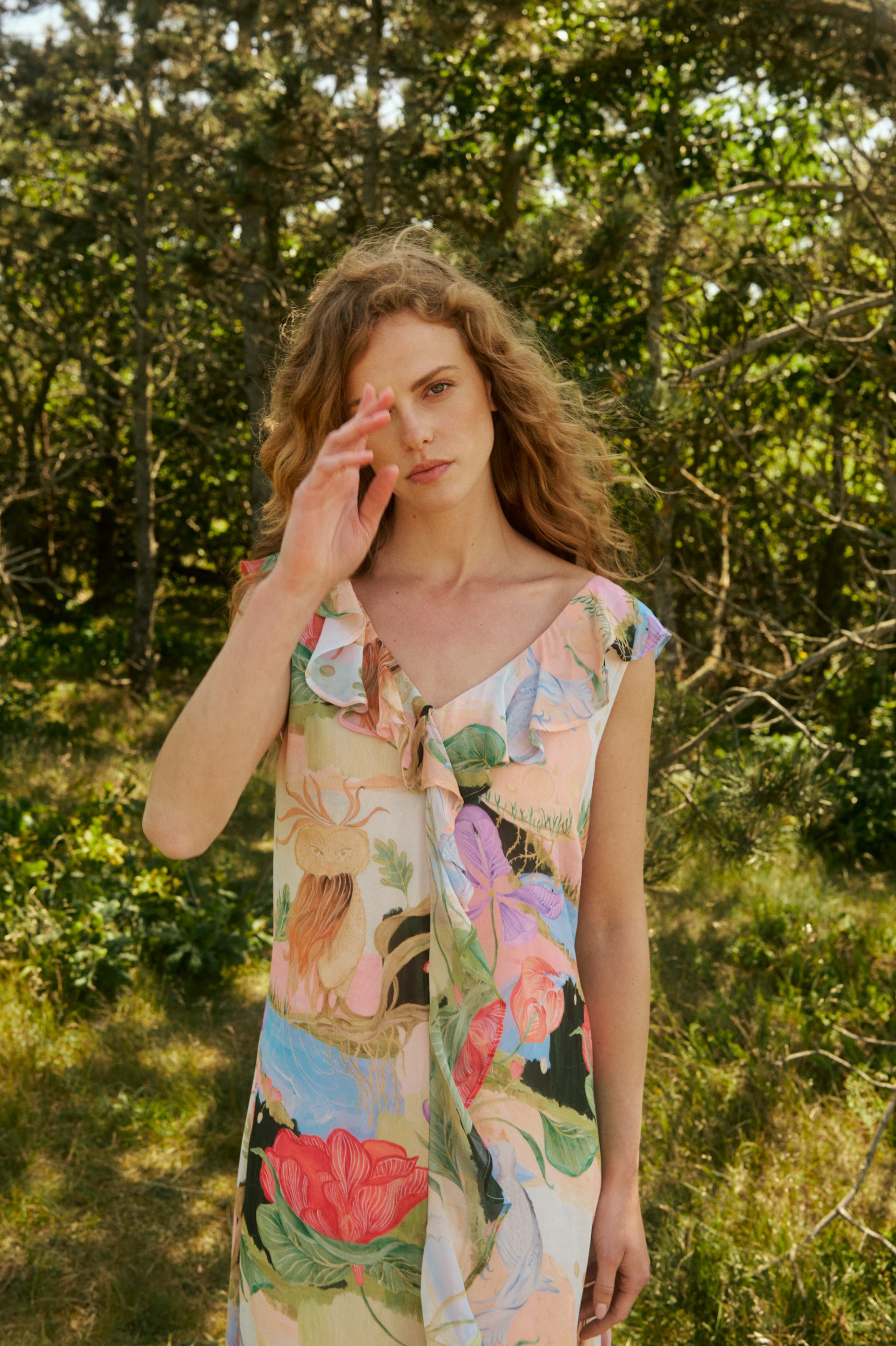 Young woman in a colorful patterned dress with a raised hand, standing in a sunny forest.