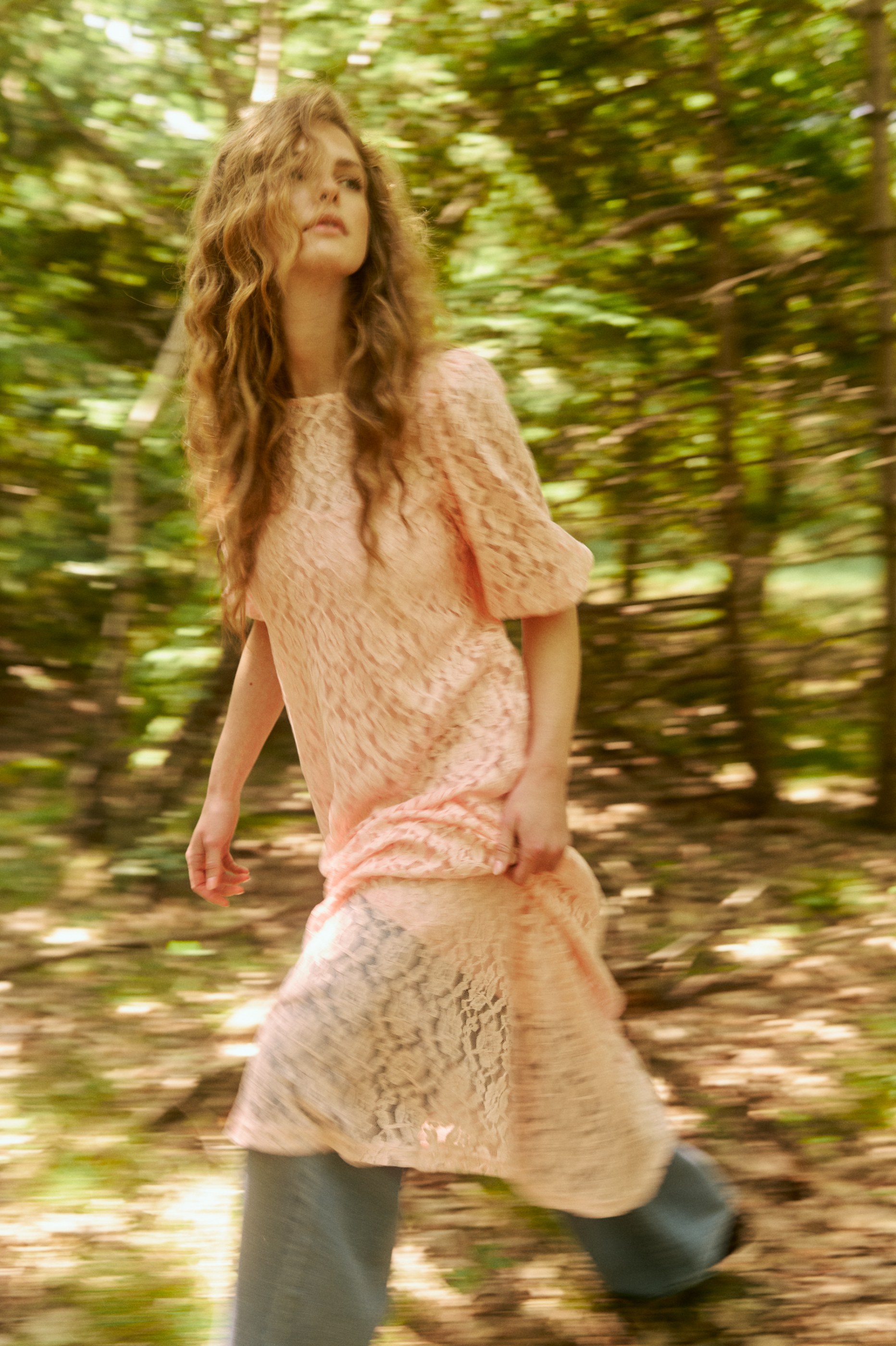Woman in pink lace dress with curly hair, walking with motion blur in a forest.