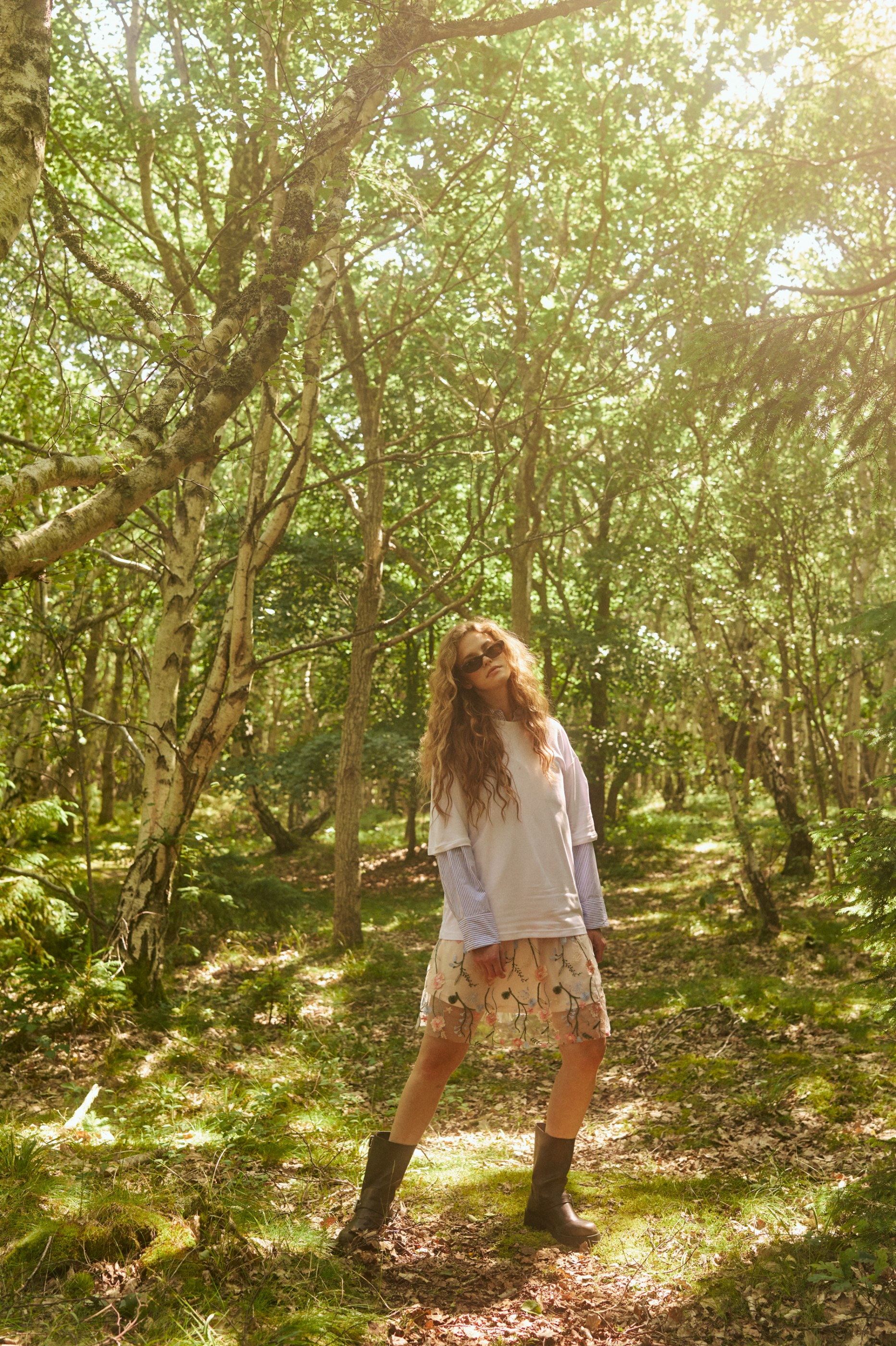 Young woman in a white tee, floral skirt, and boots stands in a dappled sunlit forest.
