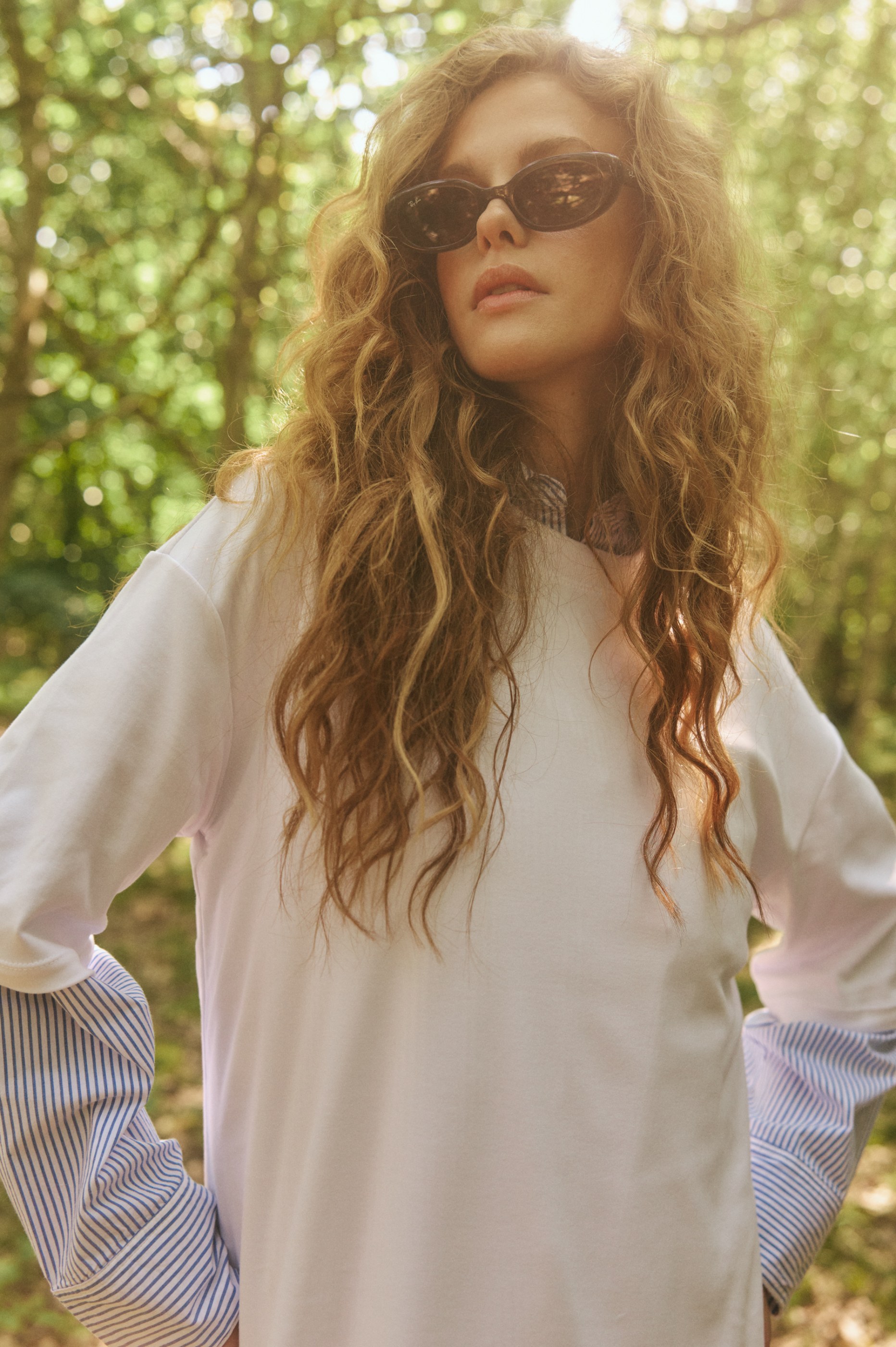 Woman with curly hair and sunglasses, white top with striped sleeves, in a sunlit natural setting.