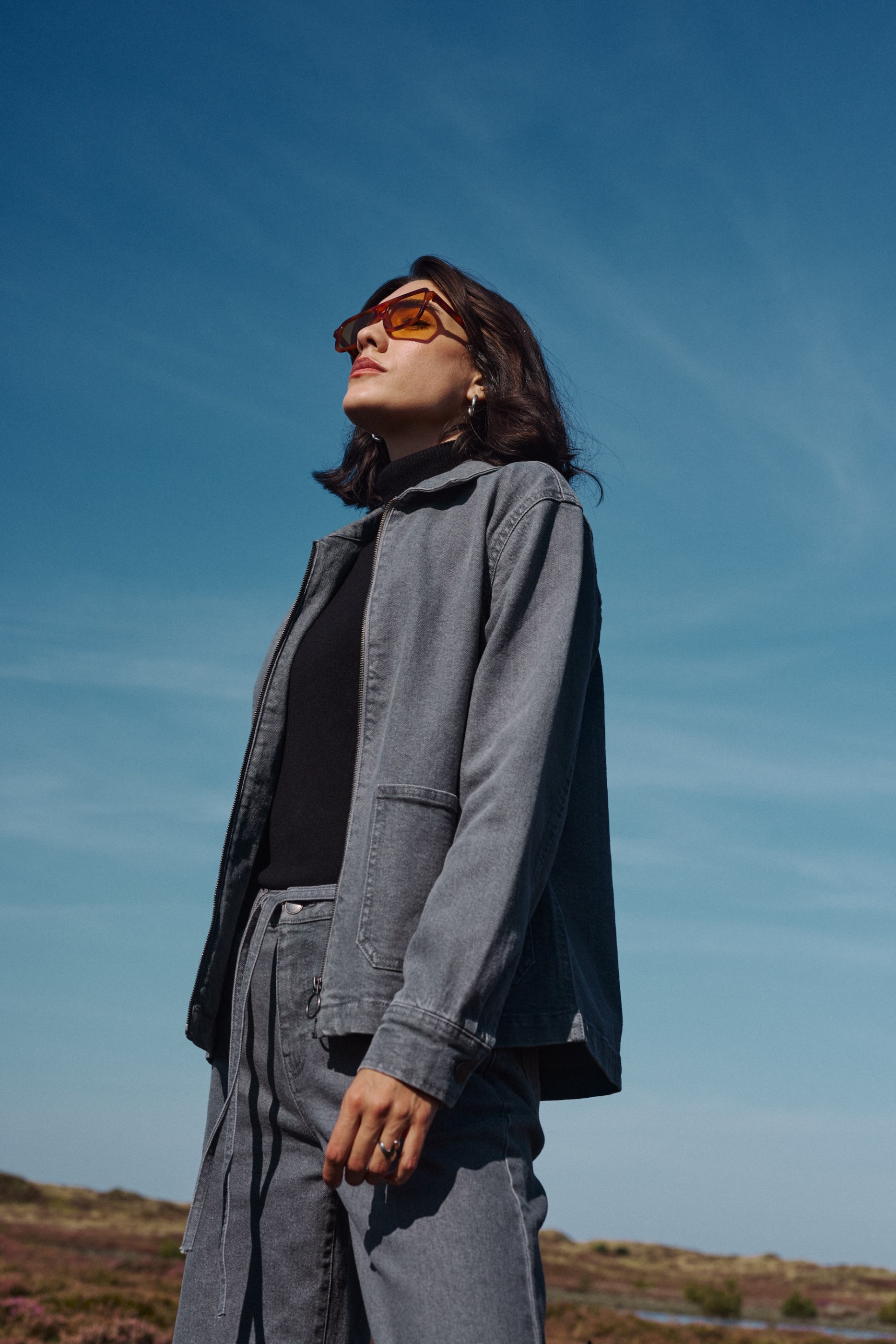 Woman in grey denim jacket and orange sunglasses looking up at a clear blue sky.