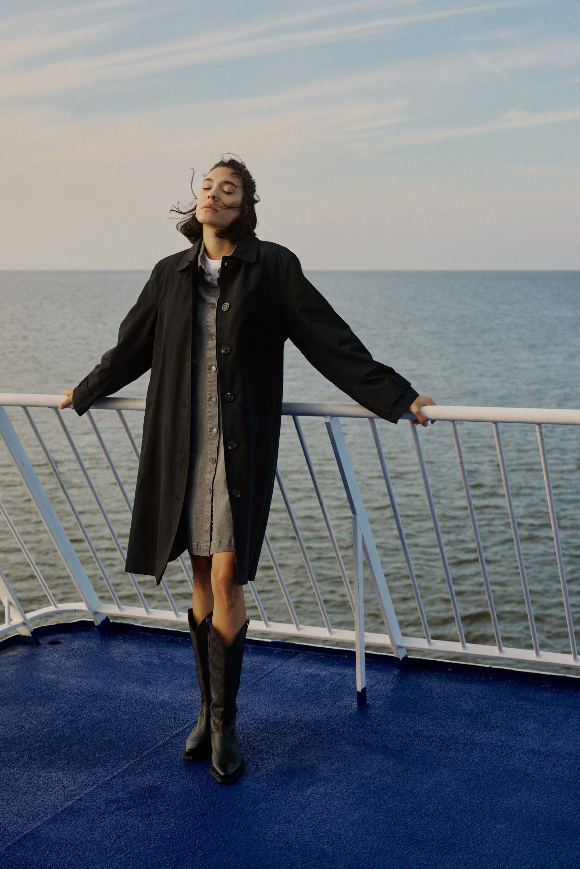 Woman in dark coat and boots, eyes closed, hair blowing on a boat deck.