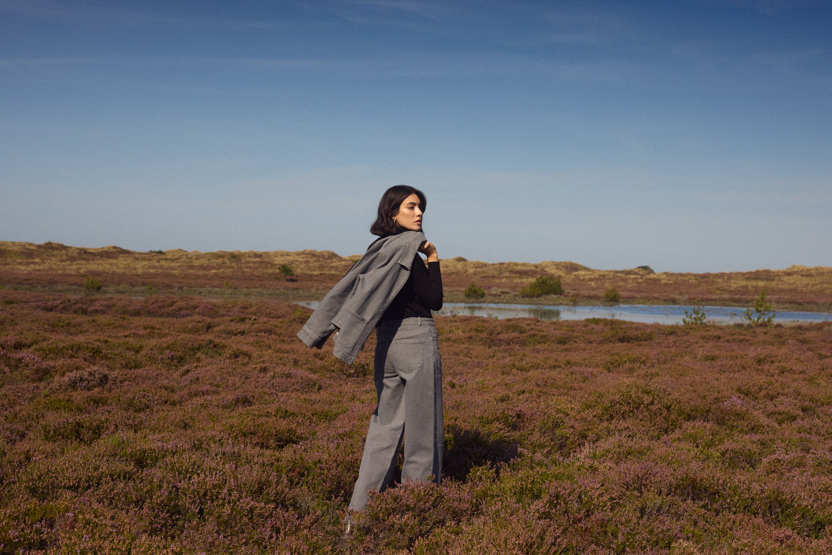 Woman in grey outfit looking over her shoulder in a purple heather field under blue sky.