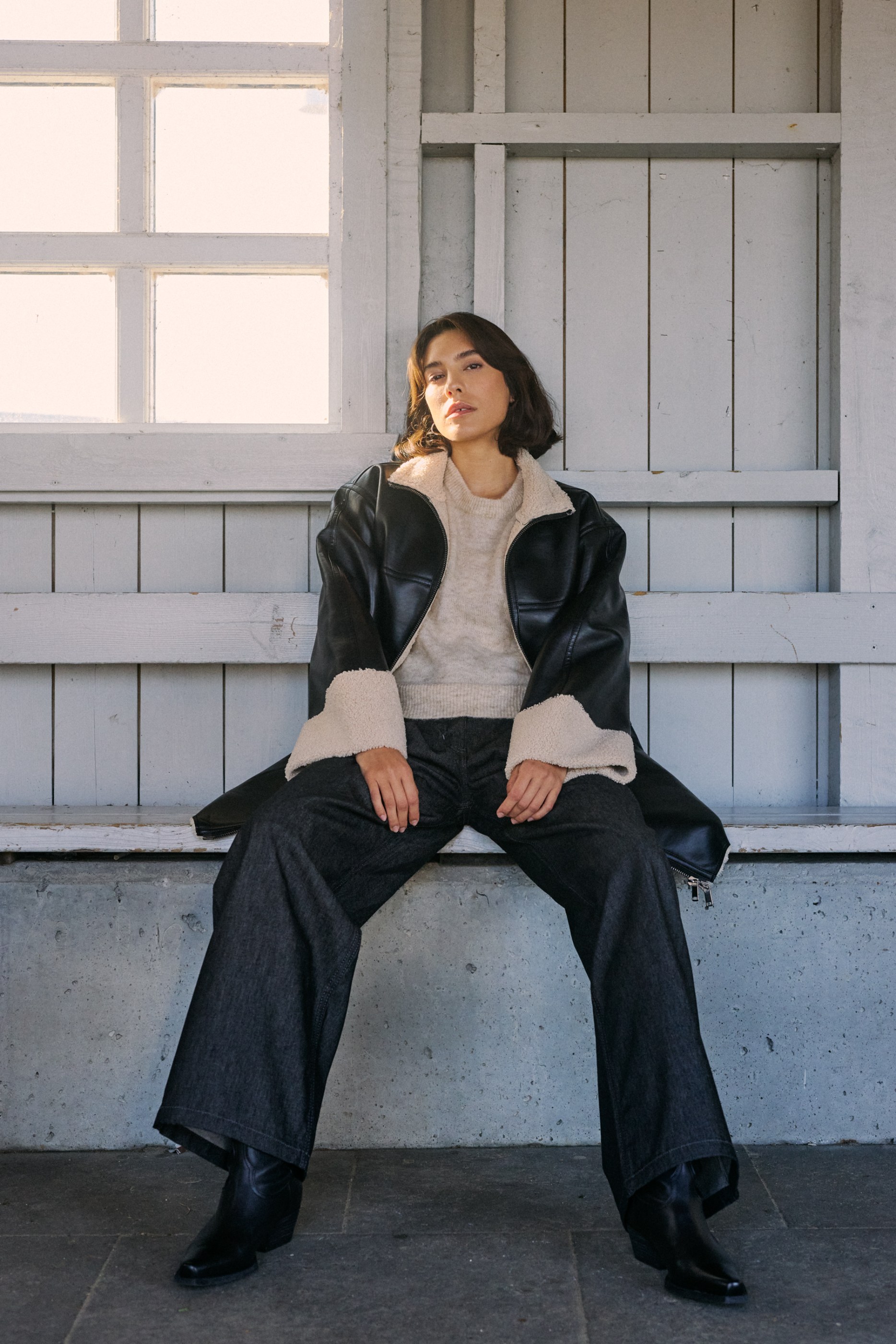 Woman with dark hair in black jacket, beige sweater, and dark jeans sitting on a concrete bench.