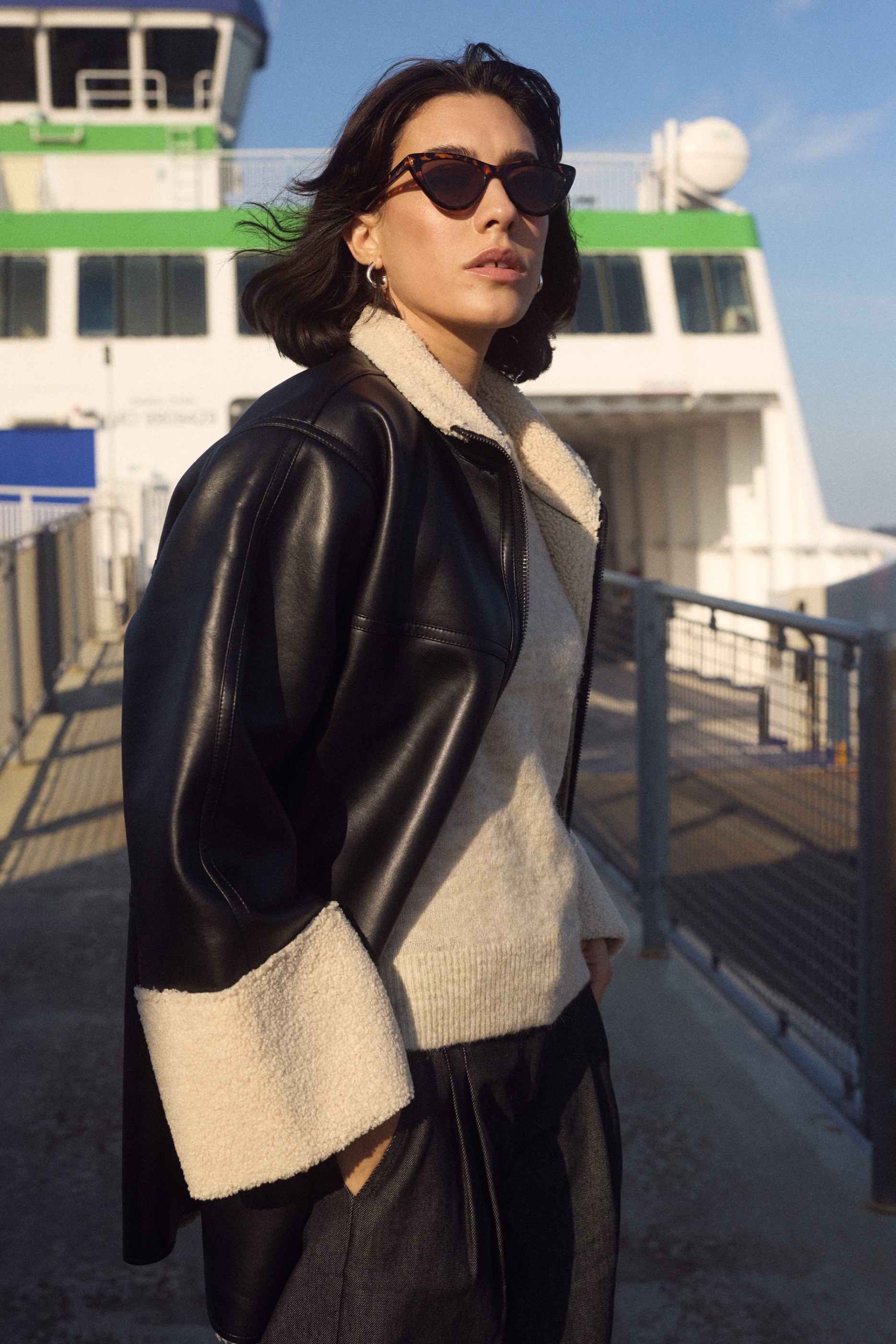 Woman in cat-eye sunglasses and a black sherpa-lined faux leather jacket by a ferry.