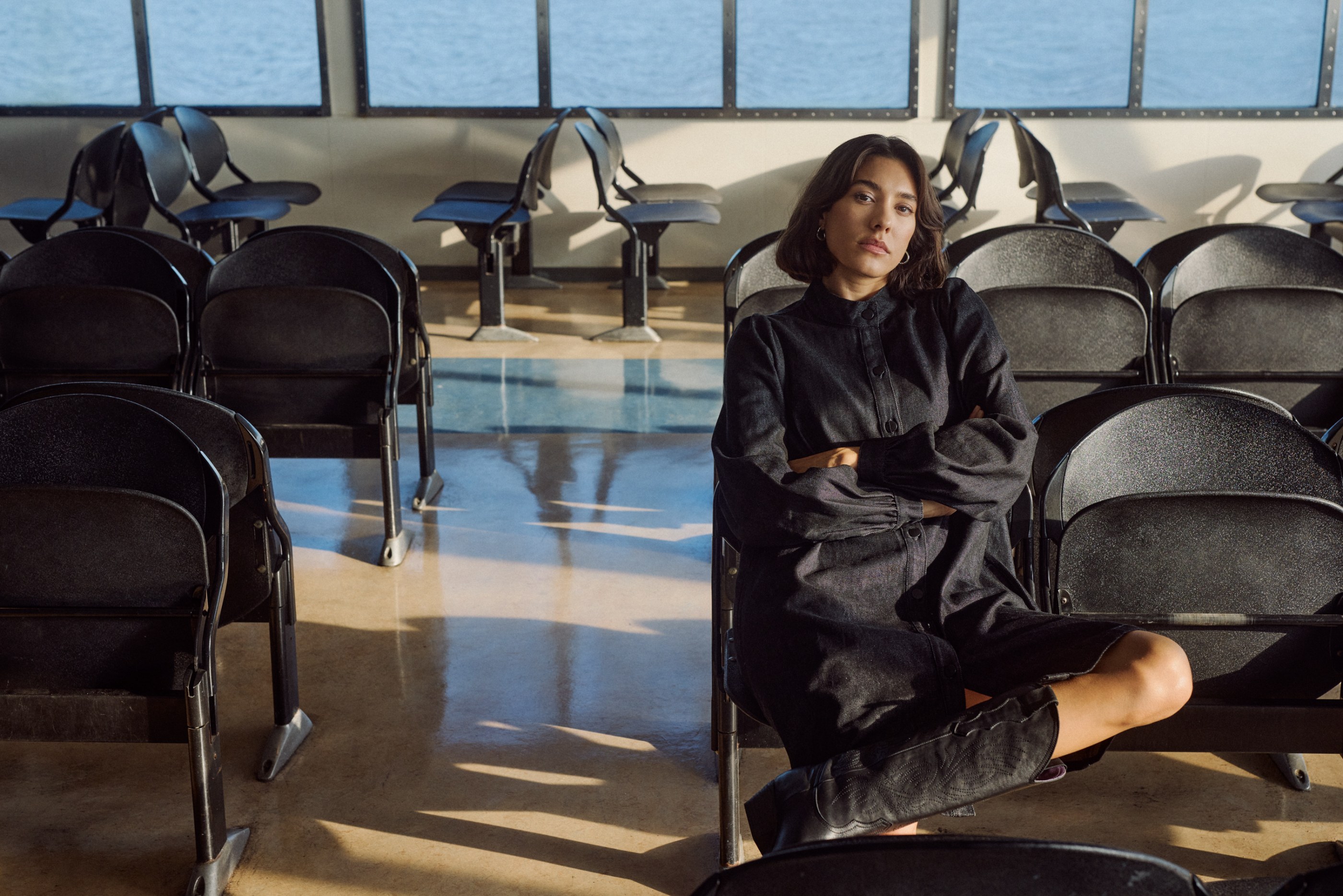 Woman in dark denim jumpsuit sitting on a ferry with arms crossed, looking at camera.