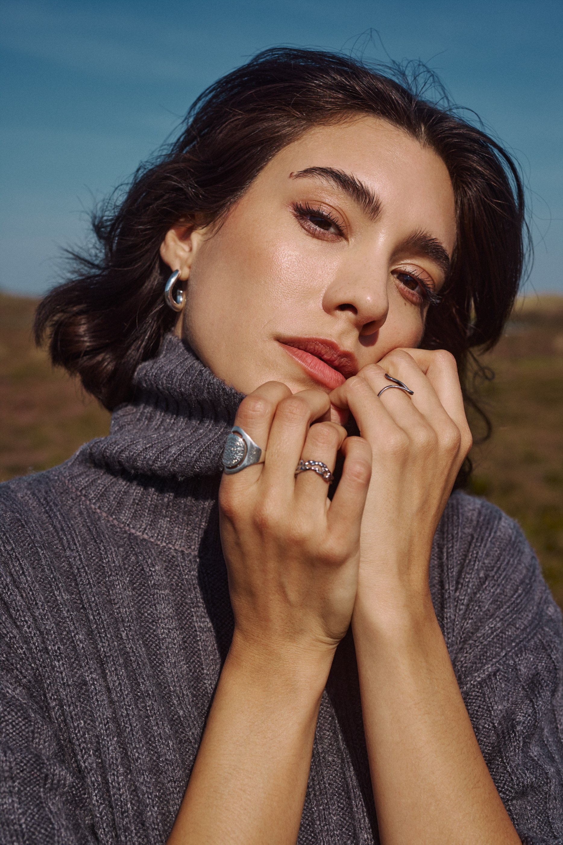 Woman in a grey turtleneck, silver jewelry, hands by face, looking at camera outdoors.